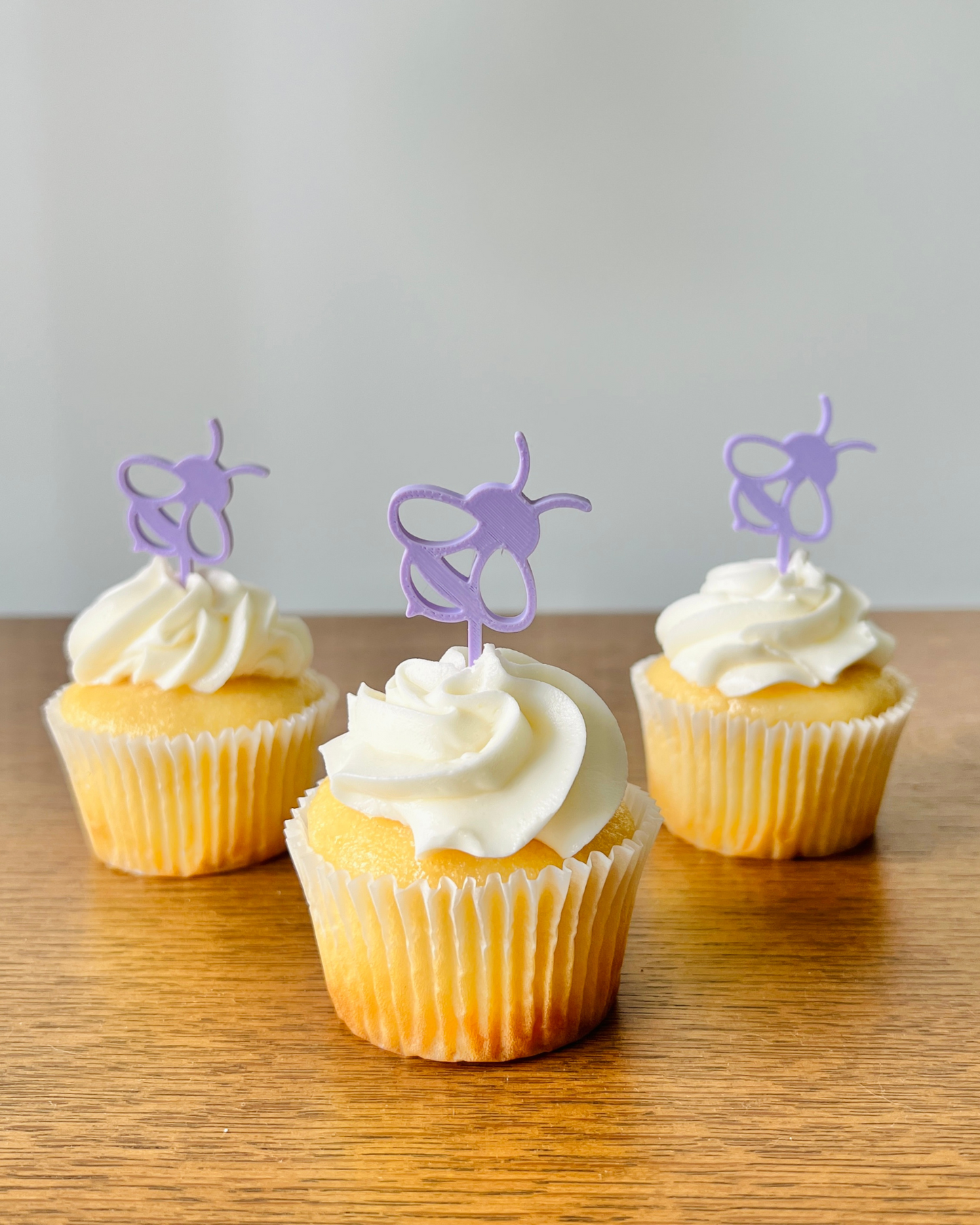 Three yellow cupcakes topped with swirls of white frosting and adorned with purple bee-shaped toppers on a wooden surface.