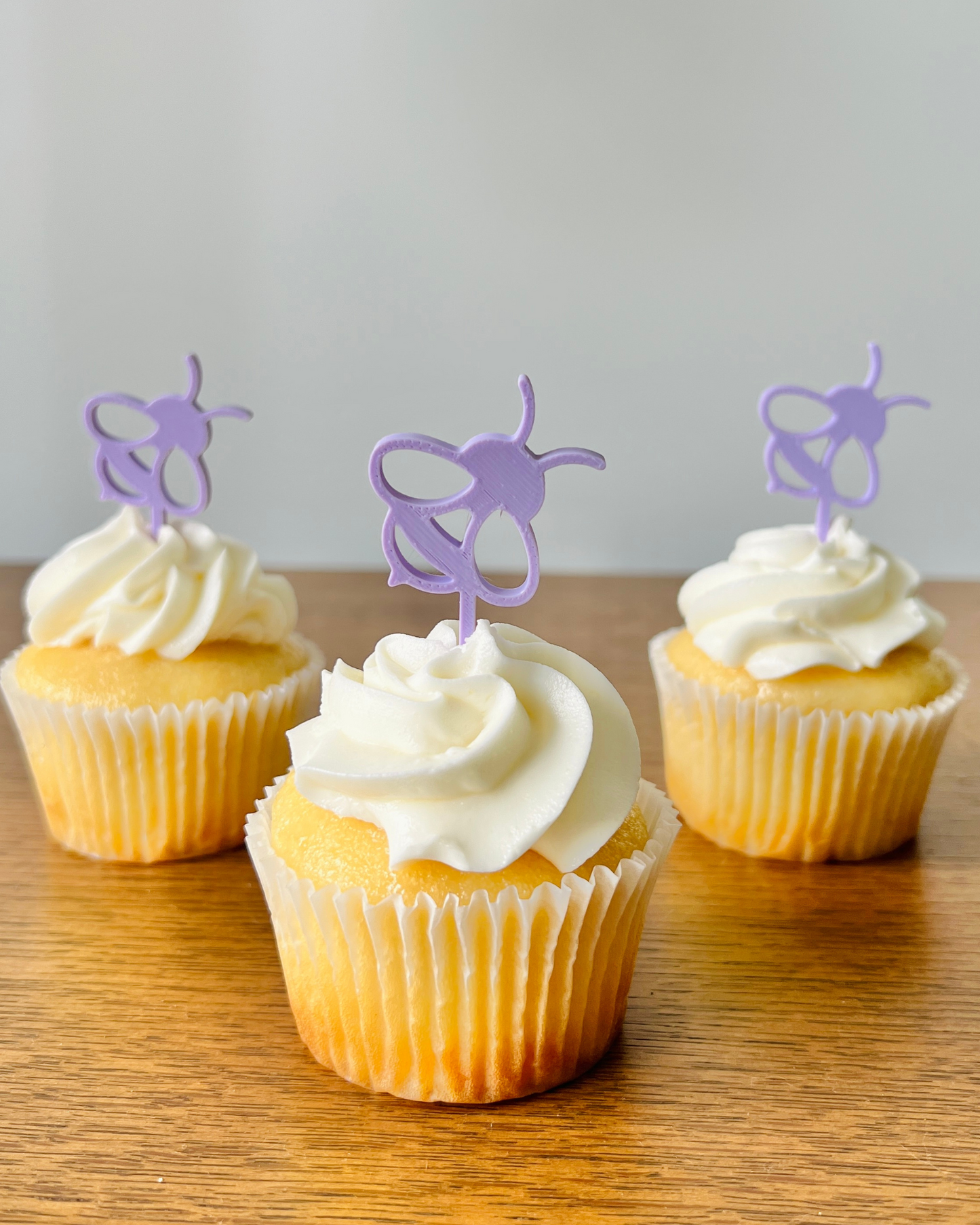 Three yellow cupcakes topped with white frosting and purple bee-shaped decorations, set on a wooden surface.