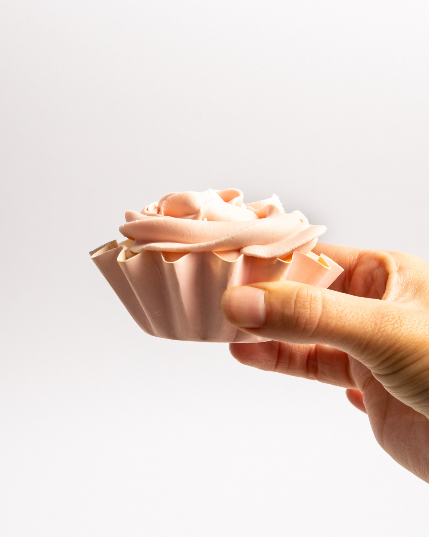 A hand holds a delicately frosted treat with soft pink icing, resembling a blooming flower against a white background.