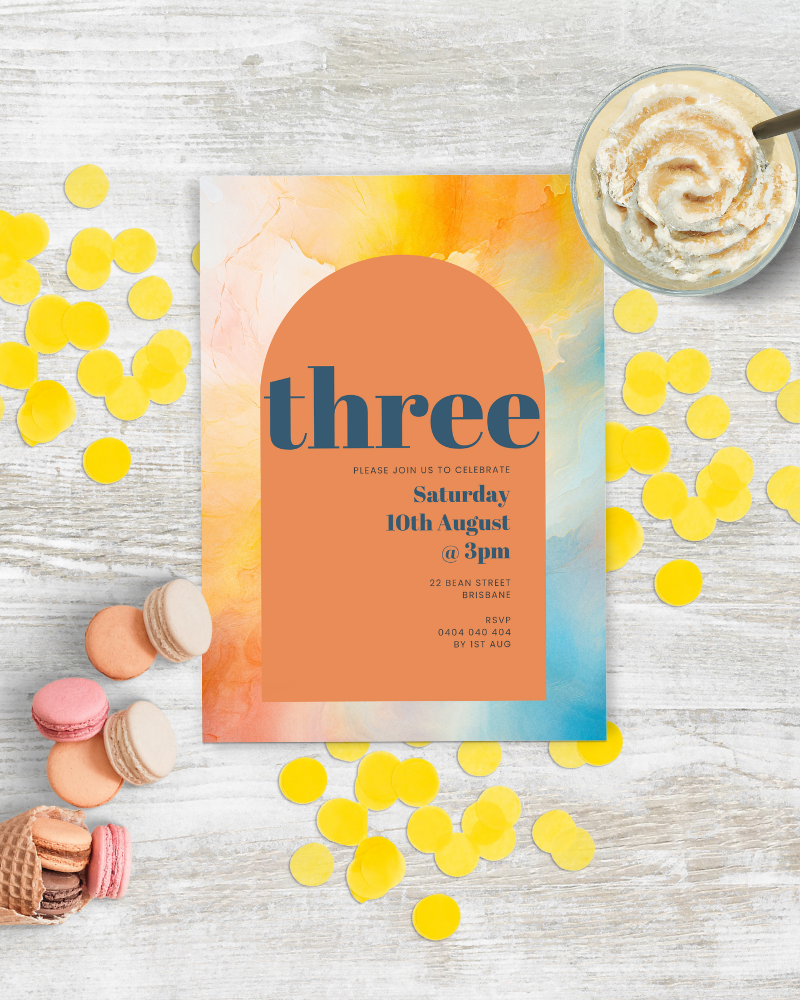 Colorful invitation with yellow confetti, macarons, and a creamy dessert on a wooden surface.