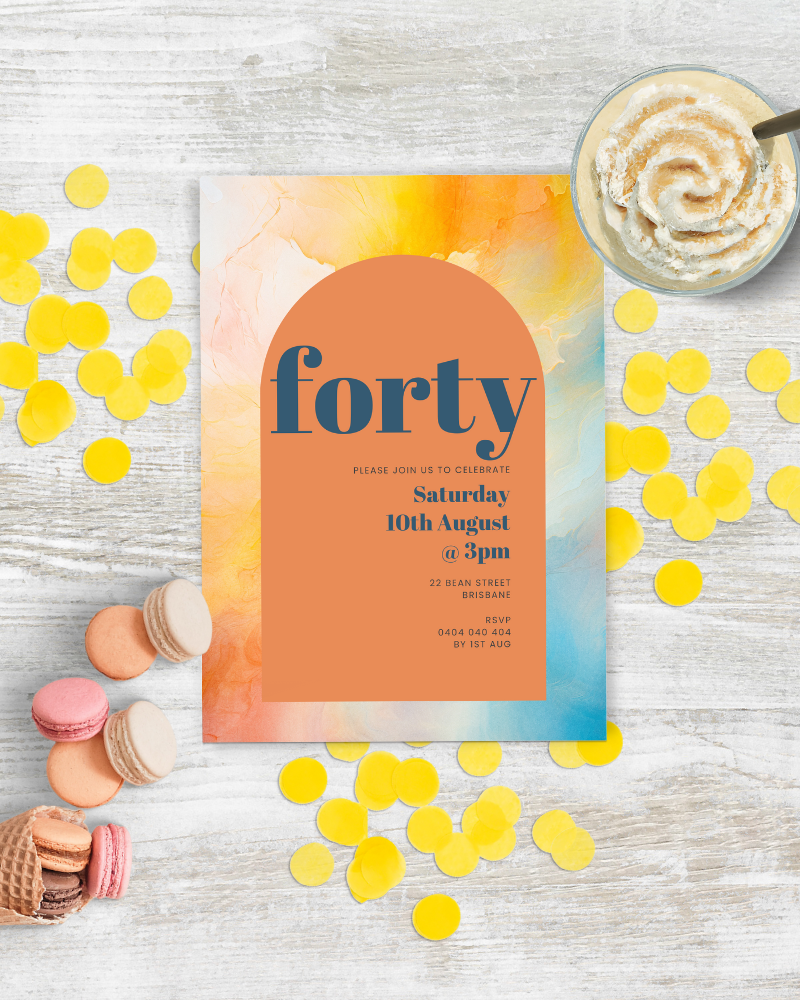 Colorful invitation on a wooden surface, surrounded by yellow confetti, macarons, and a whipped cream topped dessert.