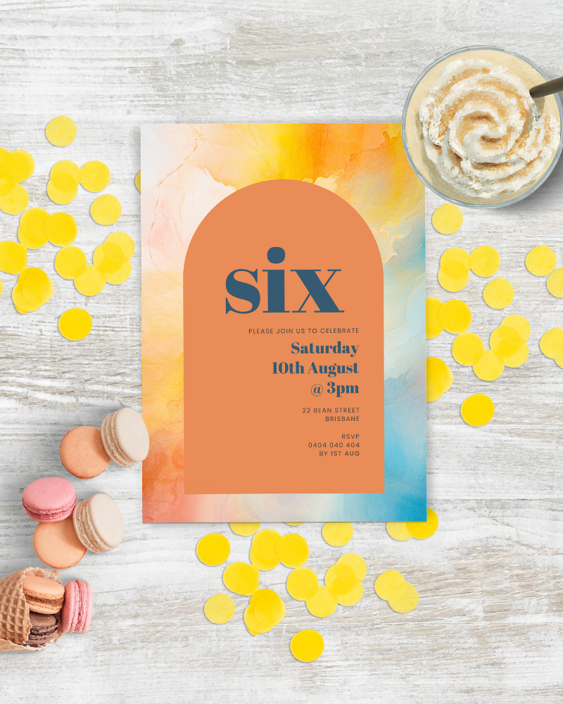 Brightly colored card announcing a celebration, surrounded by yellow confetti and sweet treats on a rustic wooden surface.