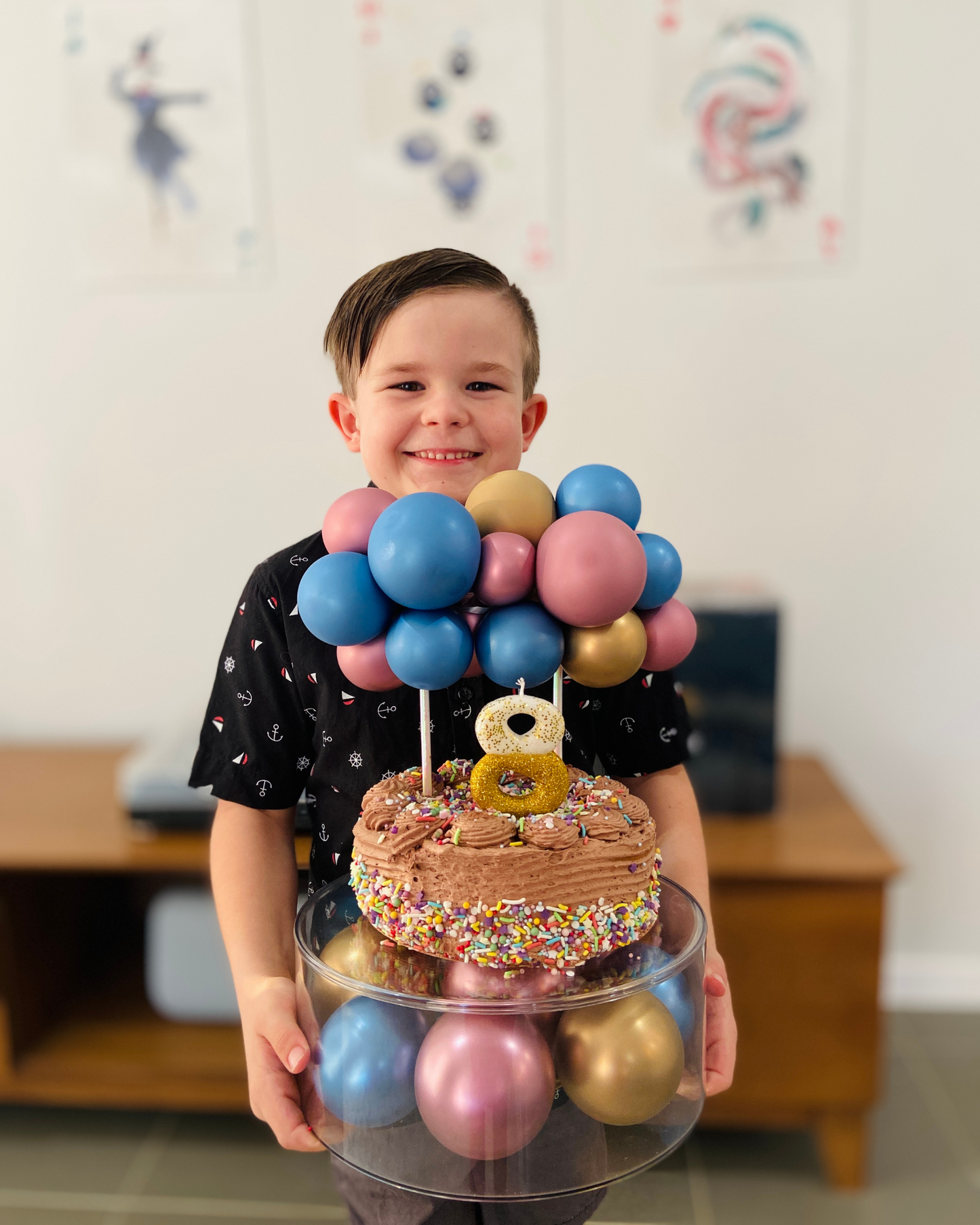 A smiling boy holds a layered cake topped with colorful balloons and a glittery number 8, celebrating a special occasion.