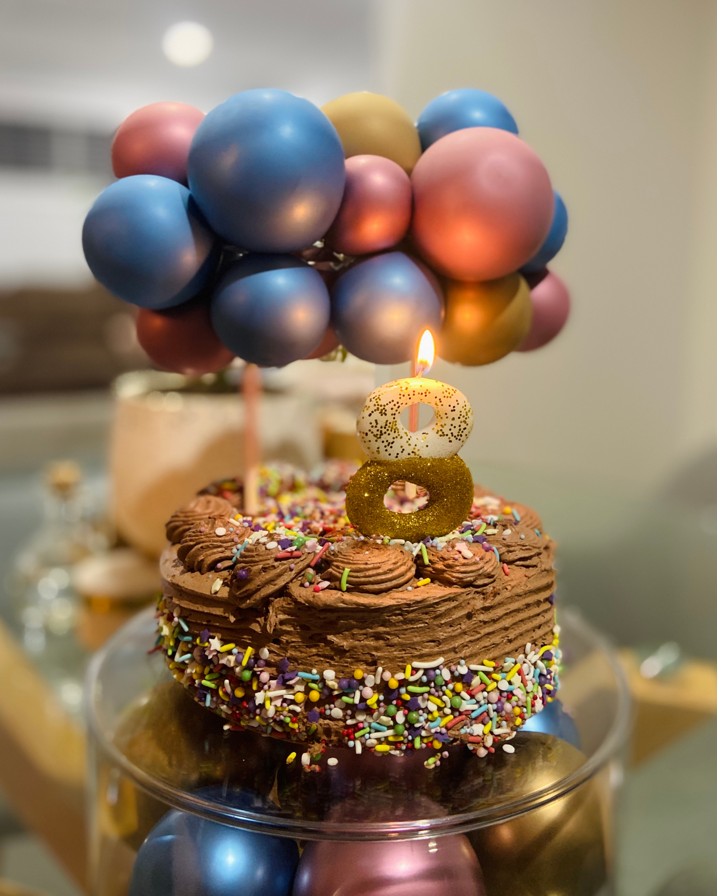 A chocolate cake topped with colorful sprinkles, a candle, and a cluster of balloons in shades of blue and pink.