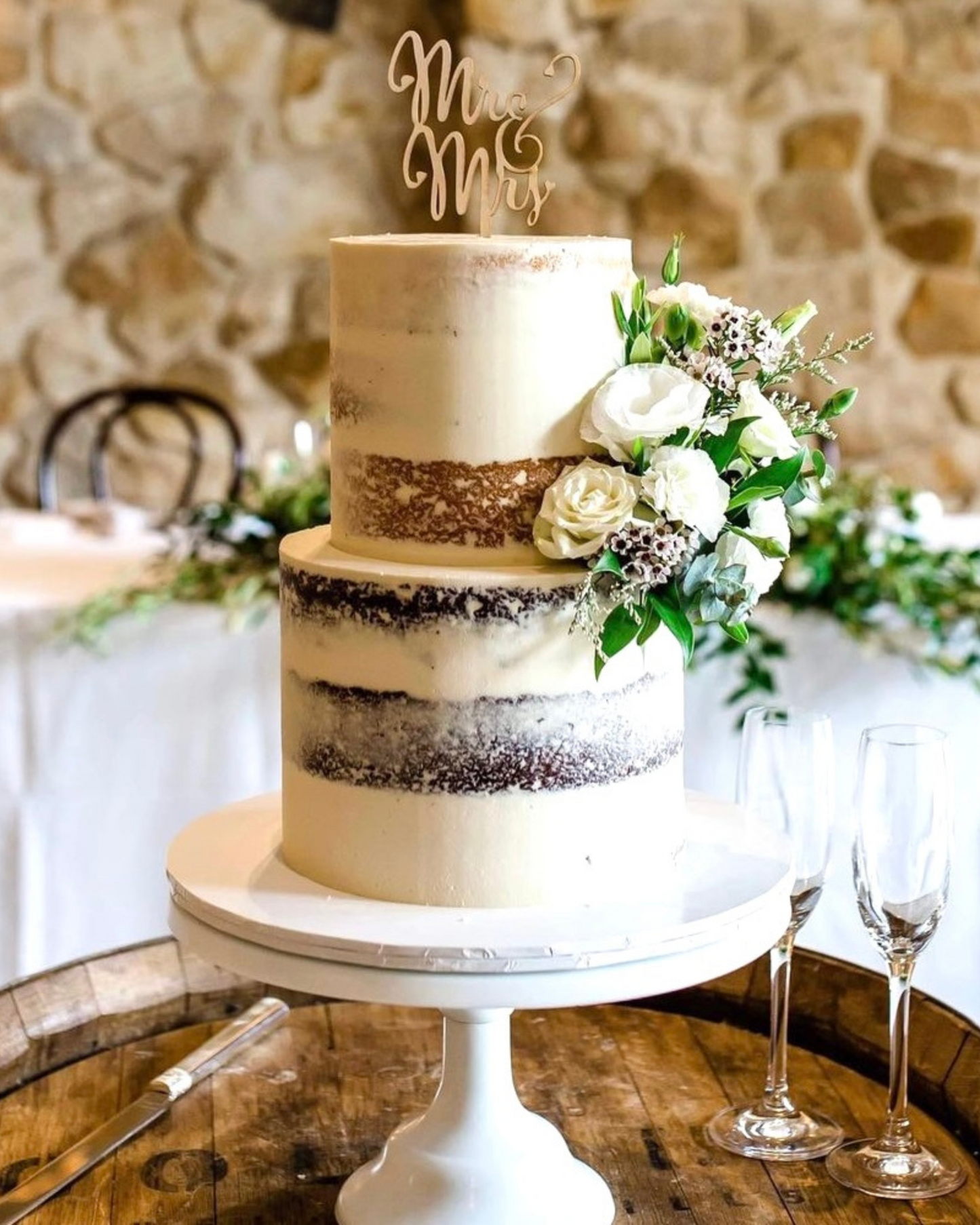 Elegant two-tier cake adorned with fresh flowers, a "Mr & Mrs" topper, set on a rustic wooden table.