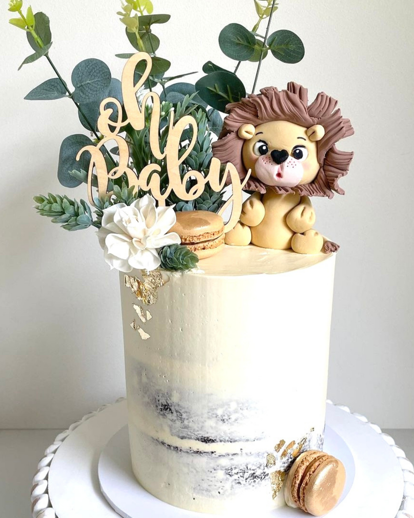 A whimsical cake topped with a lion figurine, greenery, a flower, and a playful "Oh Baby" sign.