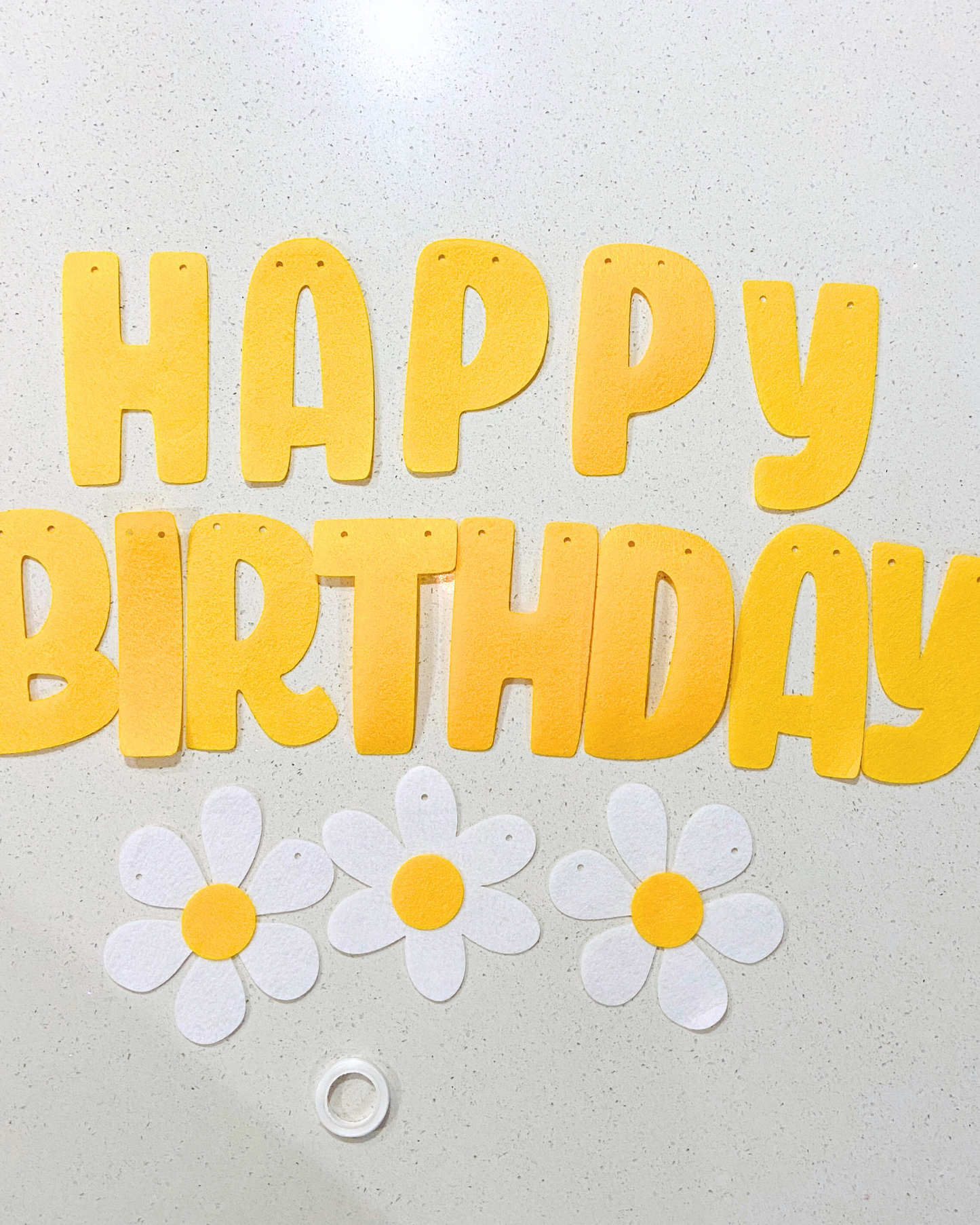 Yellow letters spell "HAPPY BIRTHDAY" alongside white flowers with yellow centers on a light countertop.