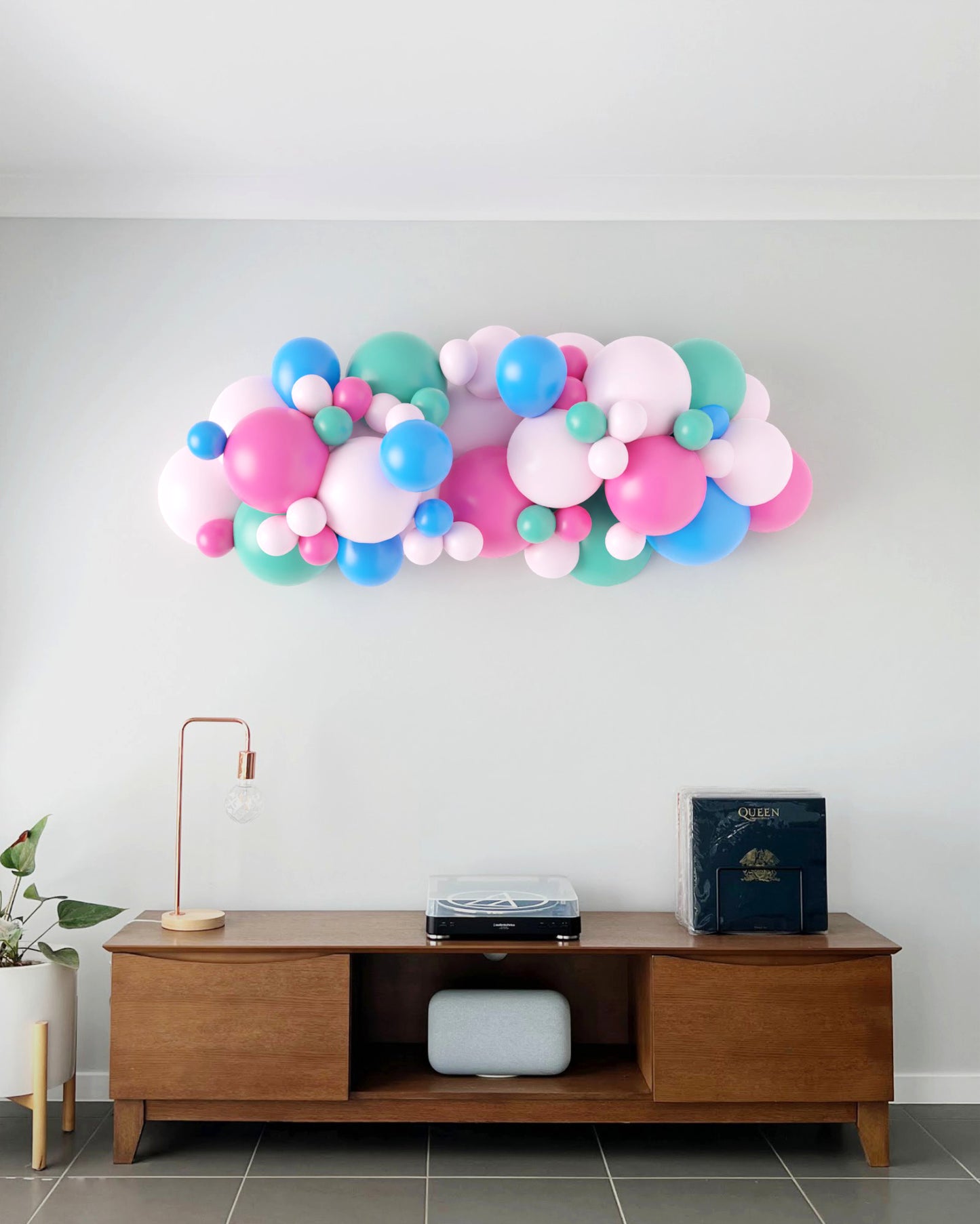 Colorful balloon garland in pink, blue, and green hues, mounted on a light wall above a wooden table and decor.
