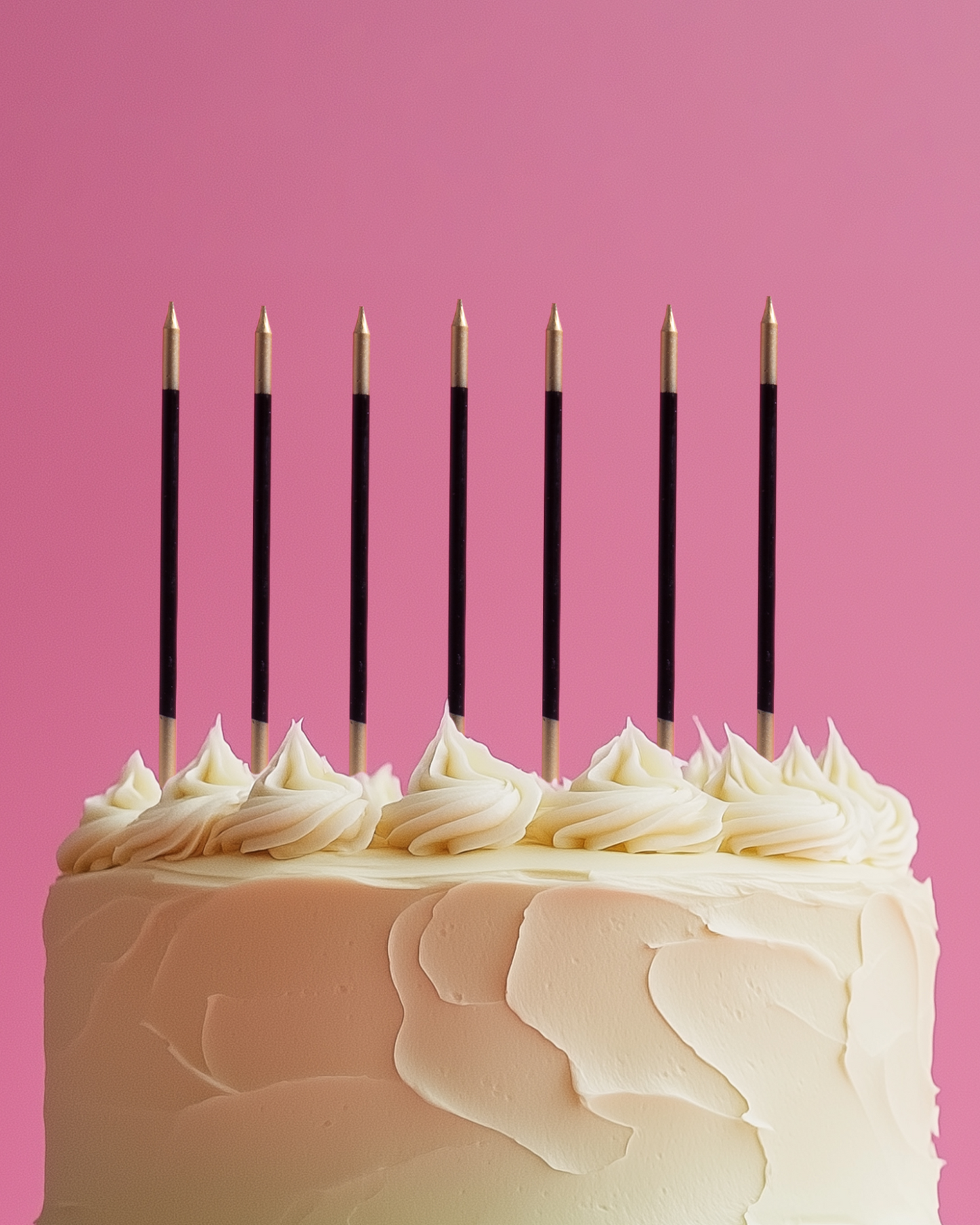 A frosted cake topped with decorative swirls and gold-tipped black candles against a pink background.
