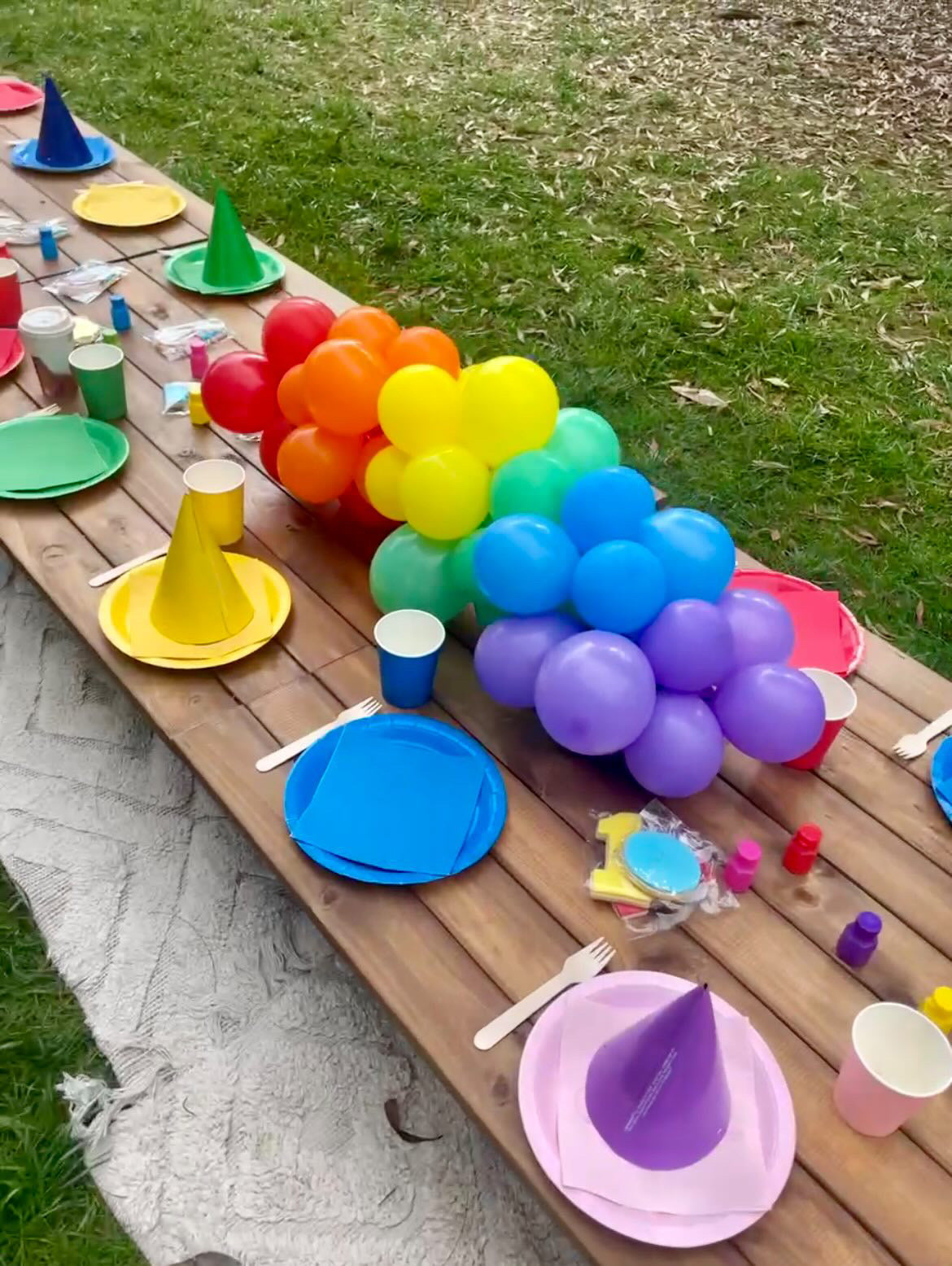 A vibrant outdoor gathering with colorful balloons, plates, cups, and party hats arranged on a wooden table.
