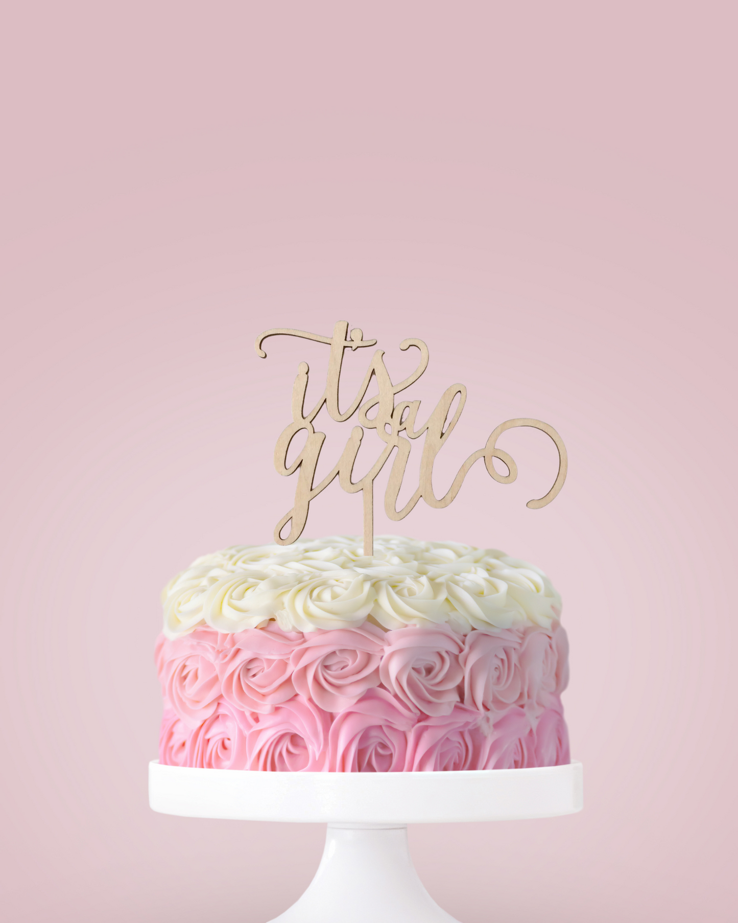 A pastel pink cake decorated with flower-like swirls, topped with a wooden "it's a girl" sign against a soft pink background.