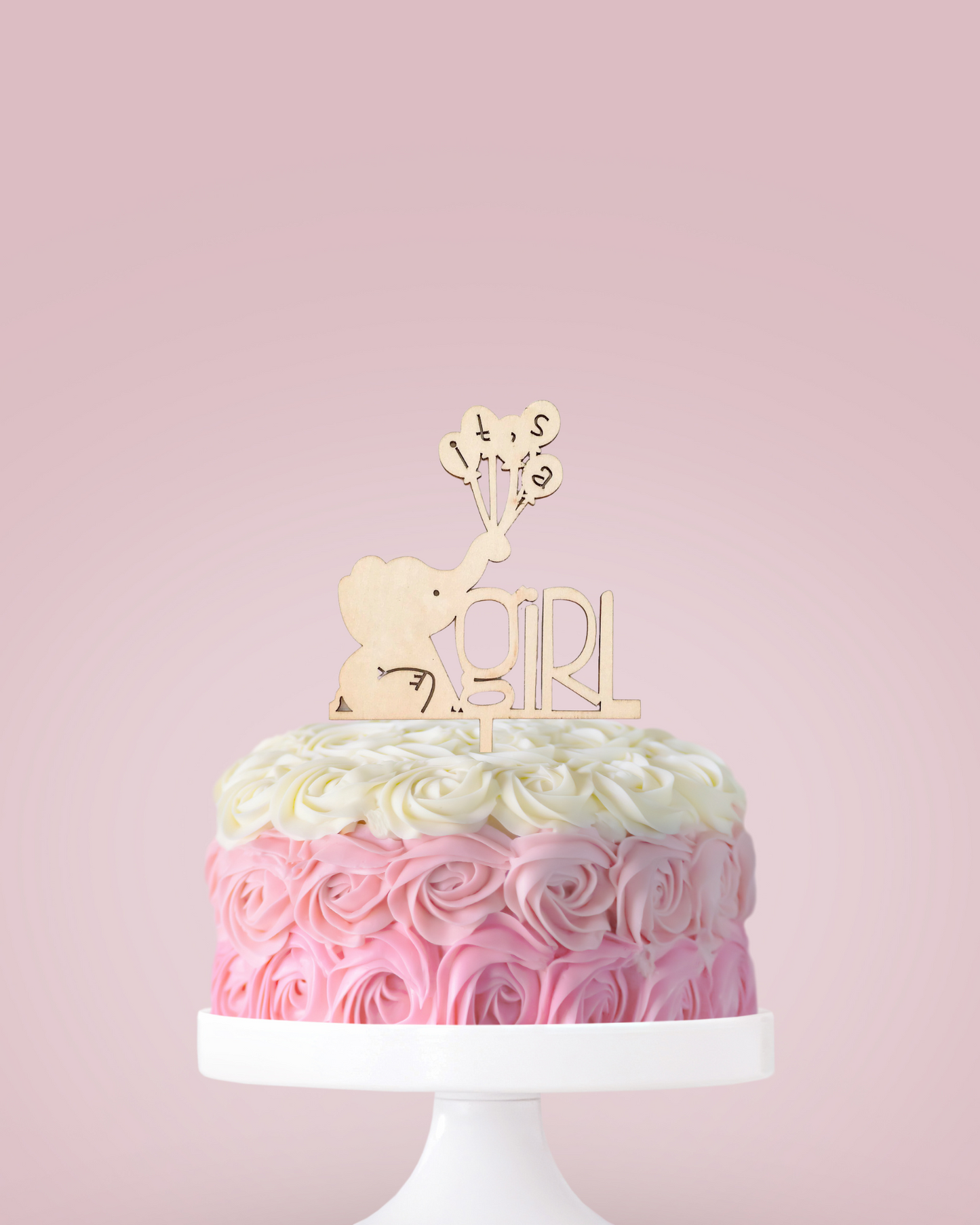 A pink and white rose-patterned cake topped with a wooden elephant and "It's a Girl" lettering, set against a soft backdrop.
