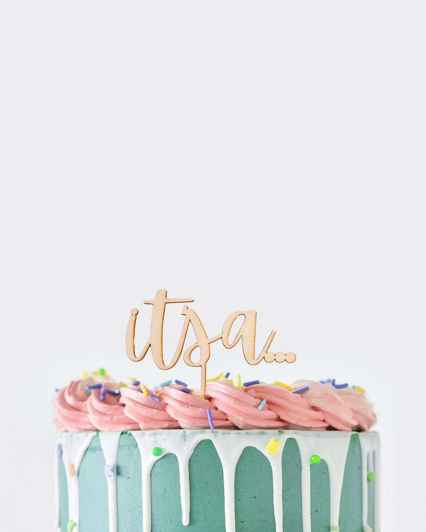 A frosted cake with pink swirls, colorful sprinkles, and a wooden topper reading "its a..." atop a turquoise base.