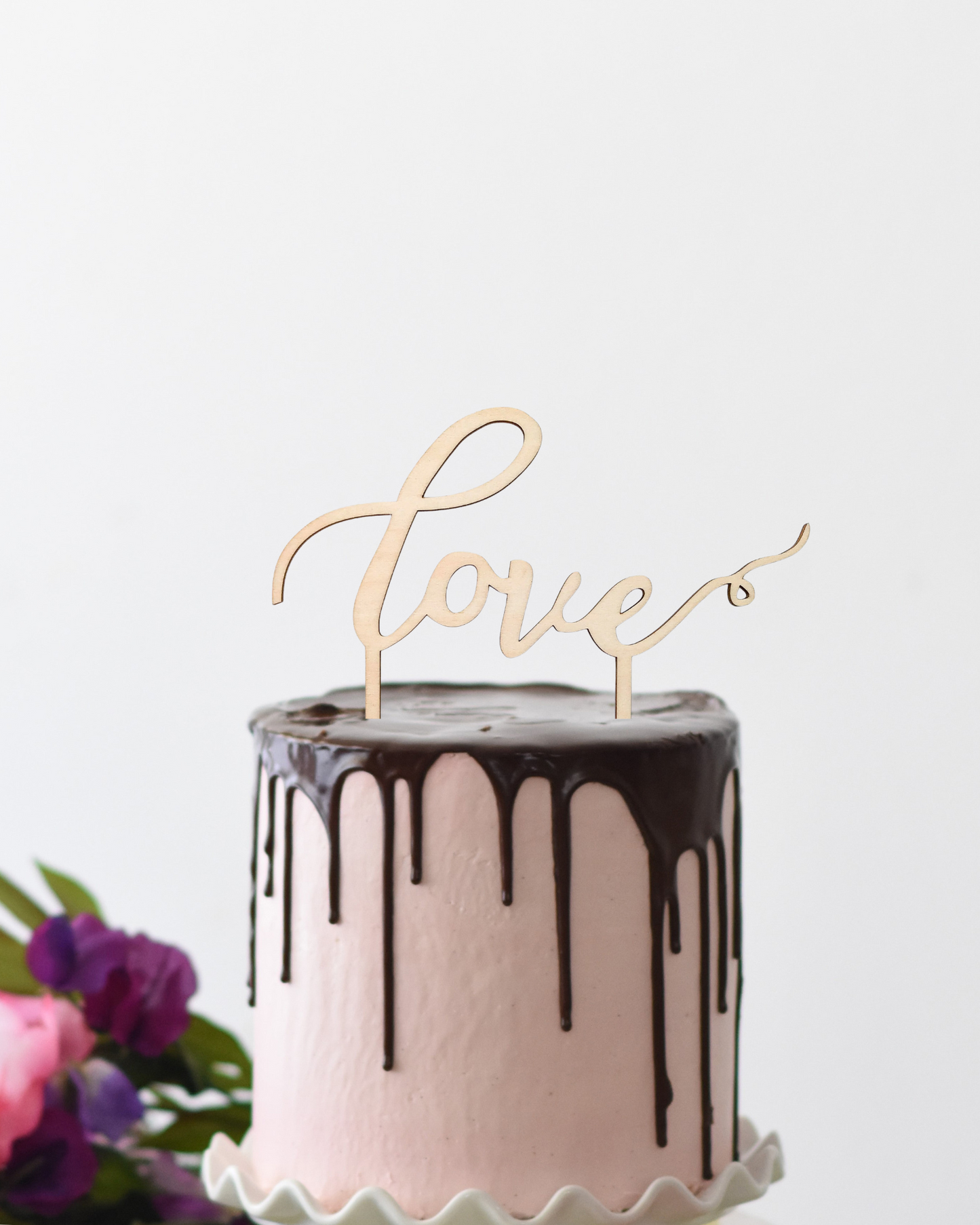 A pink cake with chocolate drizzle topped by a wooden "love" decoration and surrounded by vibrant flowers.