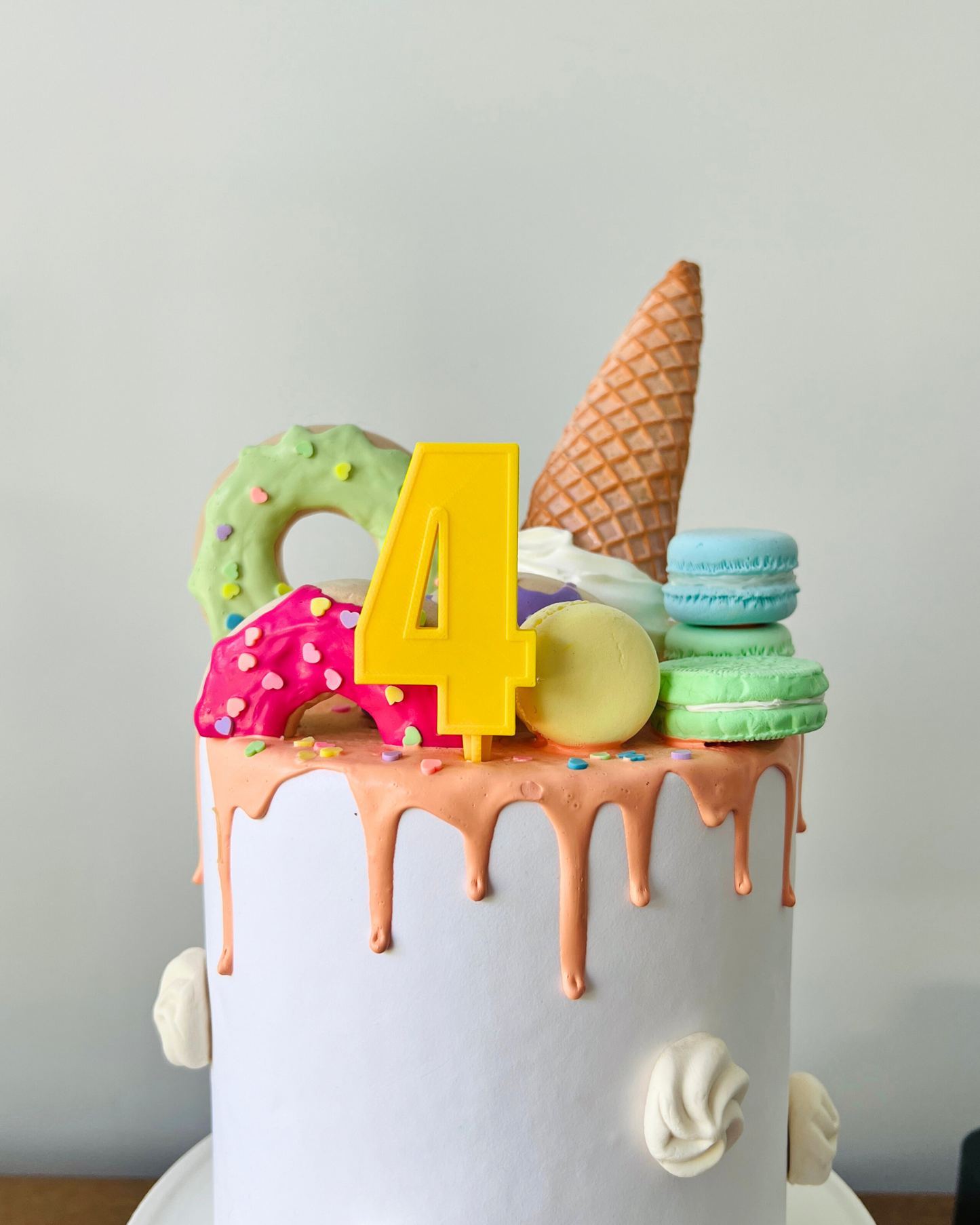 A white cake topped with colorful sweets, including a yellow number 4, macarons, and an ice cream cone.