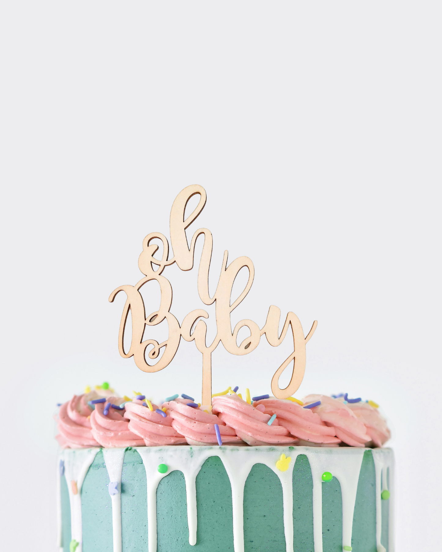 A teal cake with pink frosting, topped with a wooden "Oh Baby" topper and colorful sprinkles.