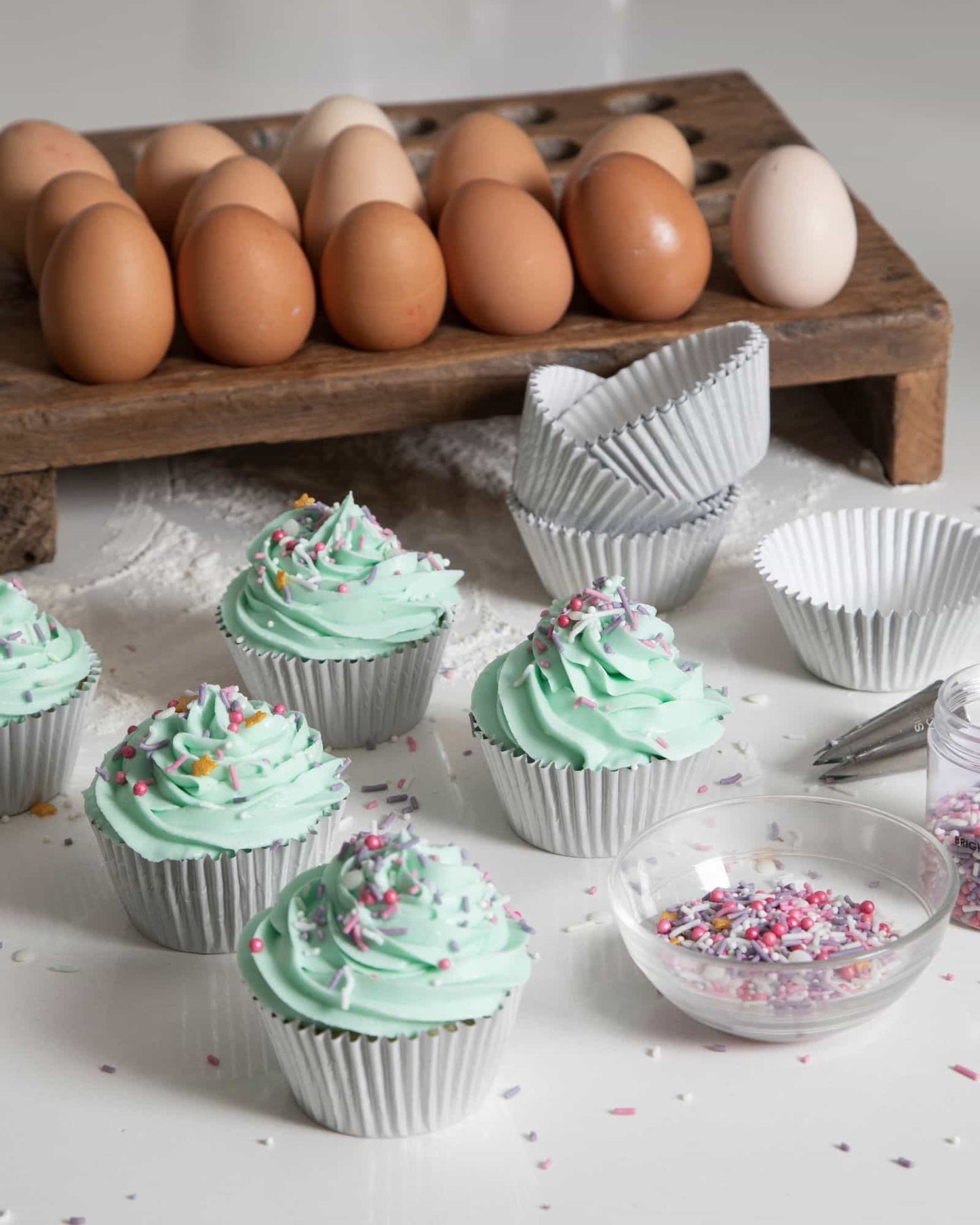 Mint green frosted treats with colorful sprinkles, alongside a wooden tray of brown and white eggs.