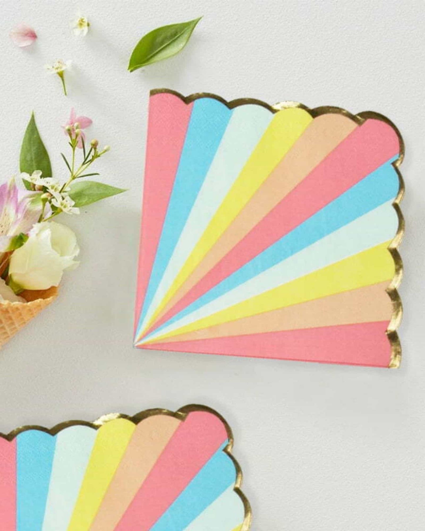Colorful fan-shaped paper with pastel rays and a gold edge, surrounded by flowers and leaves on a light surface.