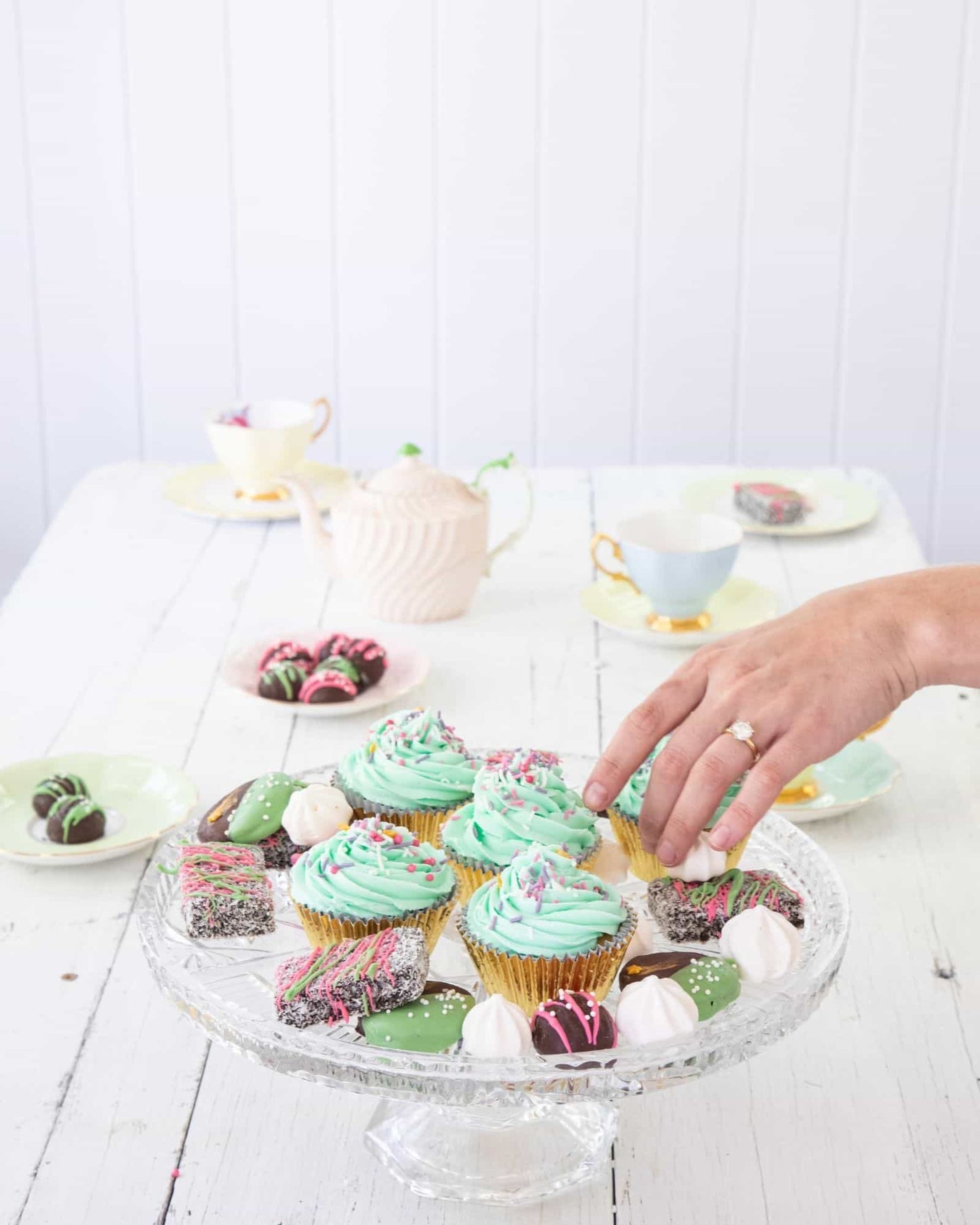 A hand reaches for colorful cupcakes and treats arranged elegantly on a glass pedestal, surrounded by delicate teacups.