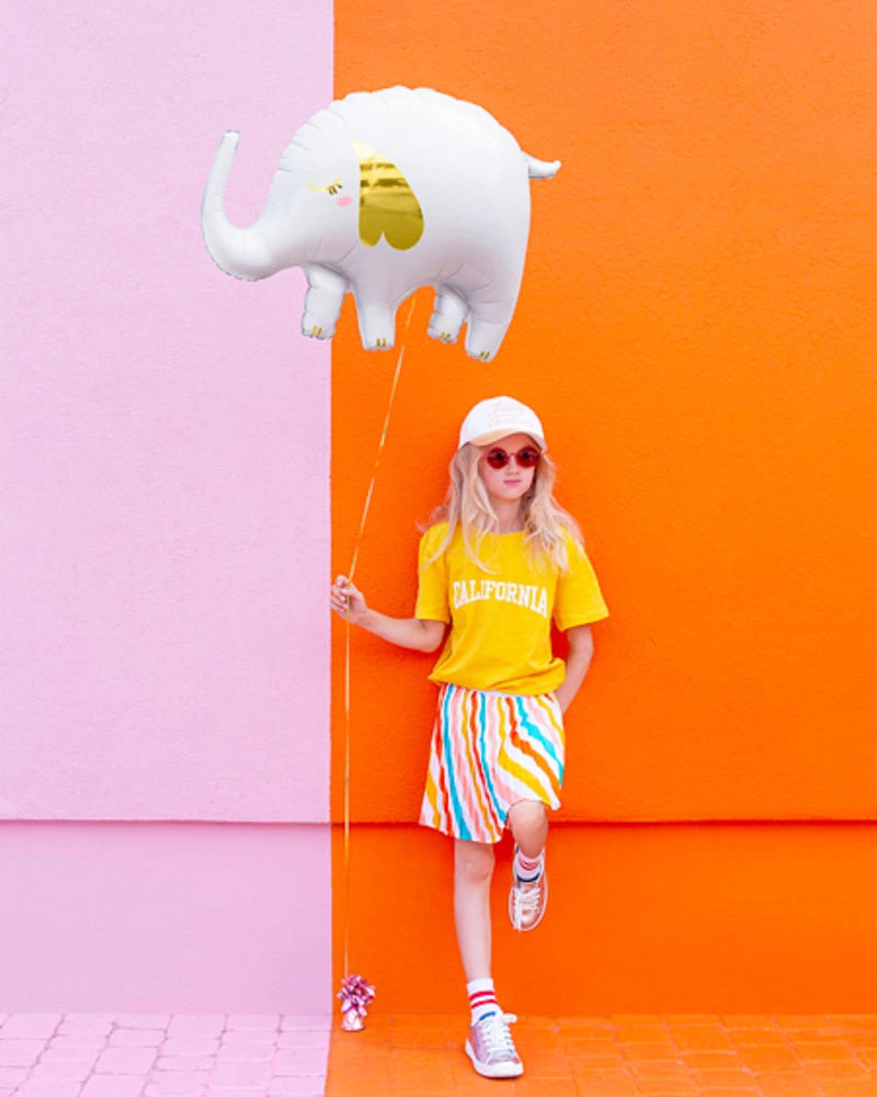 Girl in bright clothes, leaning against orange and pink wall holding a while foil elephant balloon