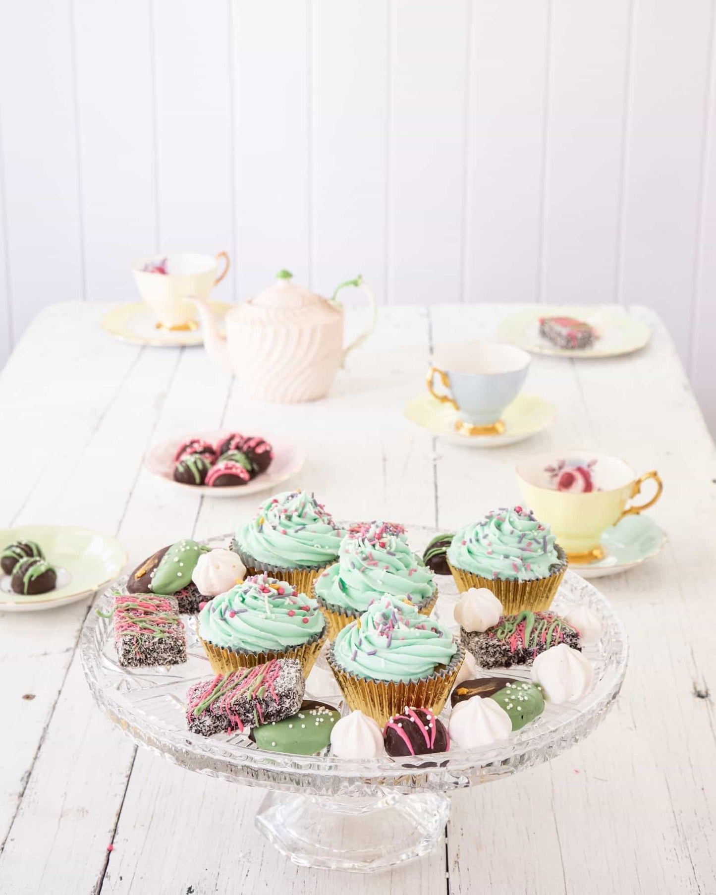 Colorful cupcakes with mint frosting and decorative sprinkles on a glass stand, surrounded by delicate tea cups and treats.