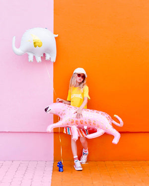 Girl in bright clothes, leaning against orange and pink wall holding a while foil elephant balloon and cuddling a pink foil leopard balloon