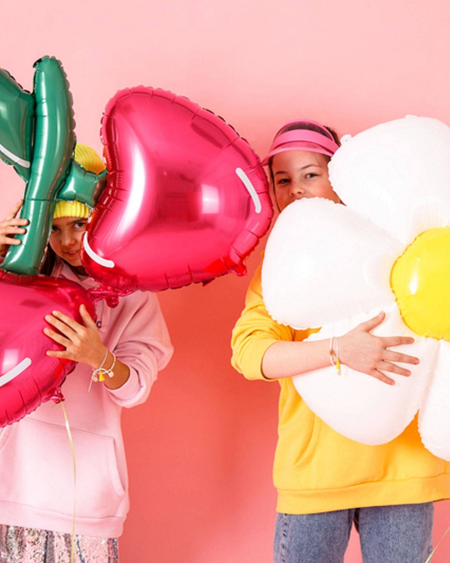 Two children hold vibrant, oversized heart and flower-shaped balloons against a soft pink background, smiling playfully.