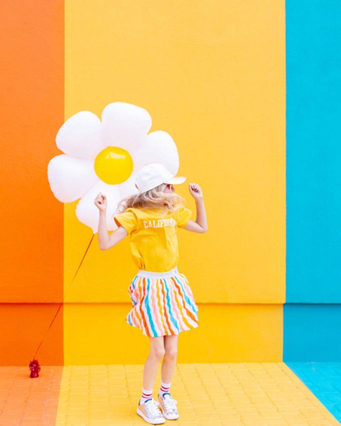 A child in a yellow shirt and striped skirt dances joyfully against a vibrant, colorful wall with a large flower balloon.