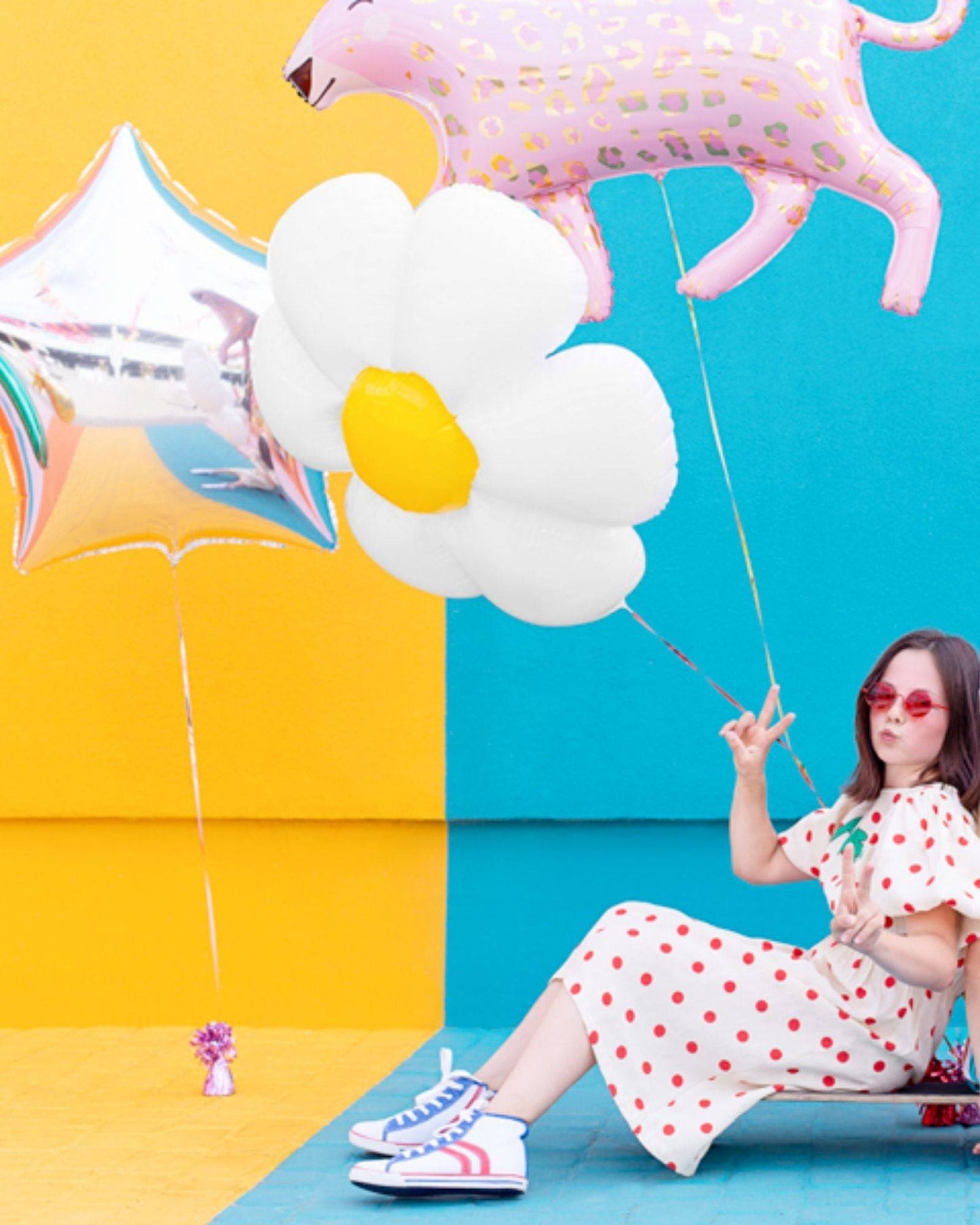 A girl in a polka dot dress poses with colorful balloons against a vibrant yellow and blue backdrop, wearing stylish sneakers
