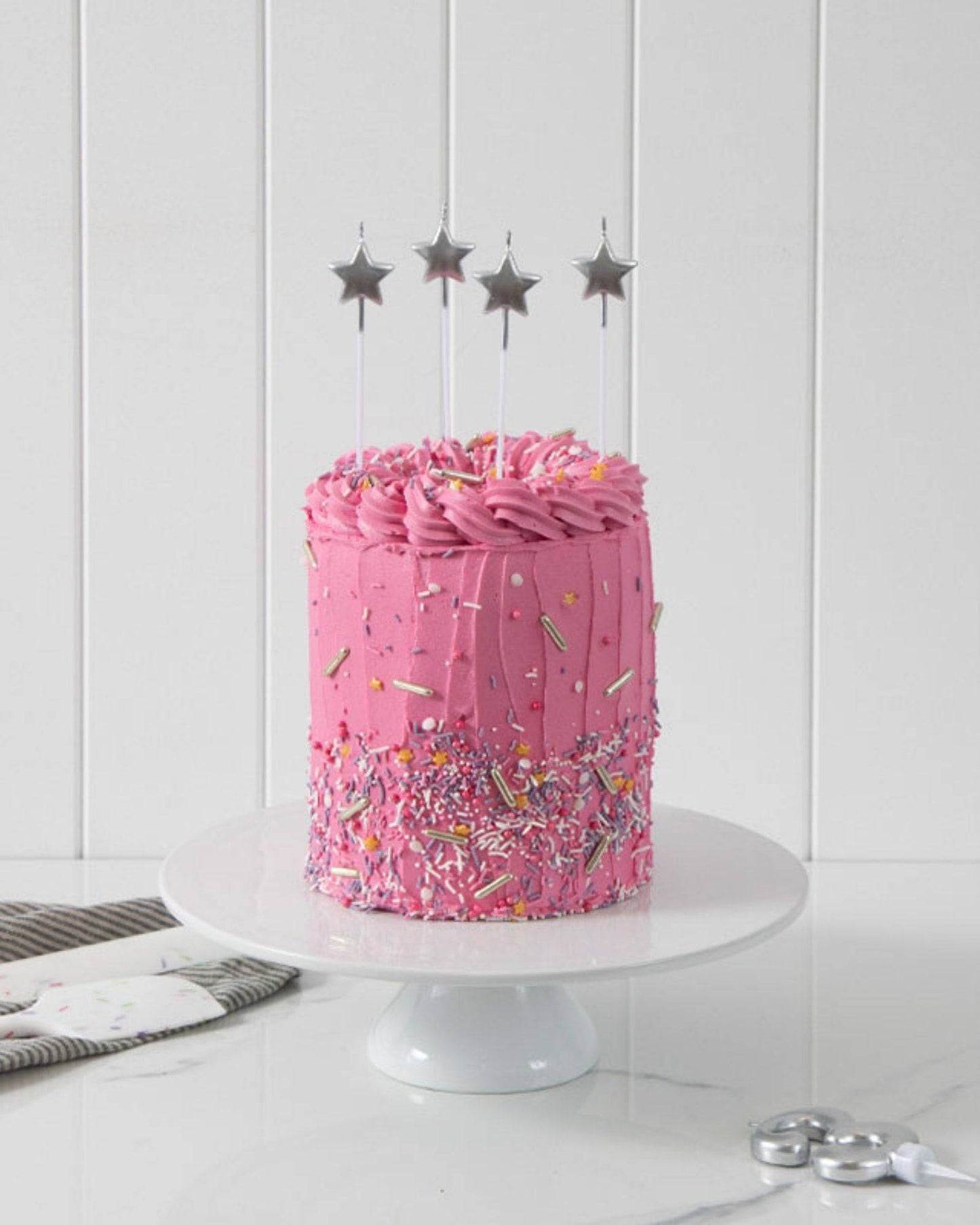 A pink frosted cake adorned with colorful sprinkles and topped with silver star candles on a white pedestal.