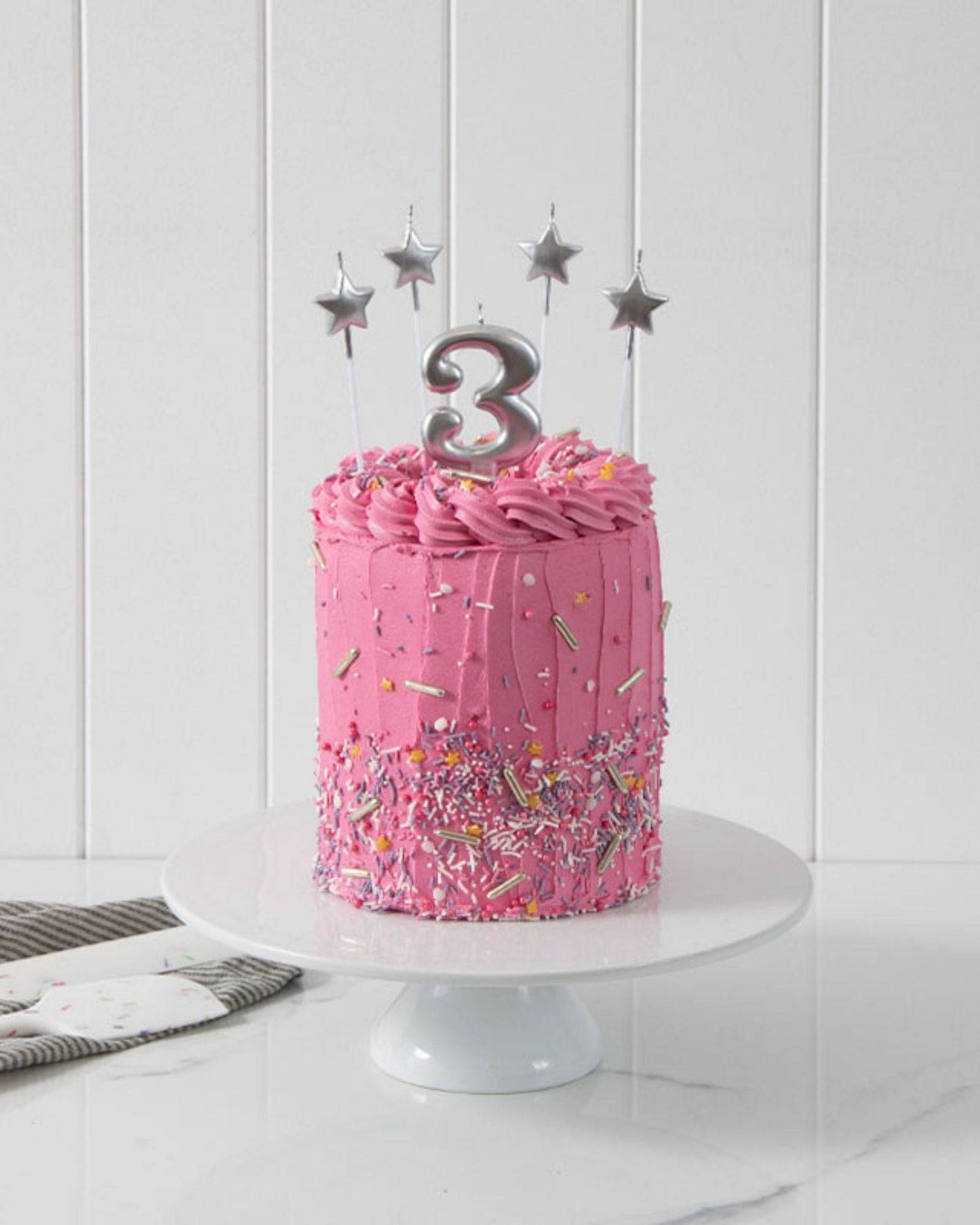 A pink layered cake with swirls, topped with a silver "3" and star candles, surrounded by colorful sprinkles.