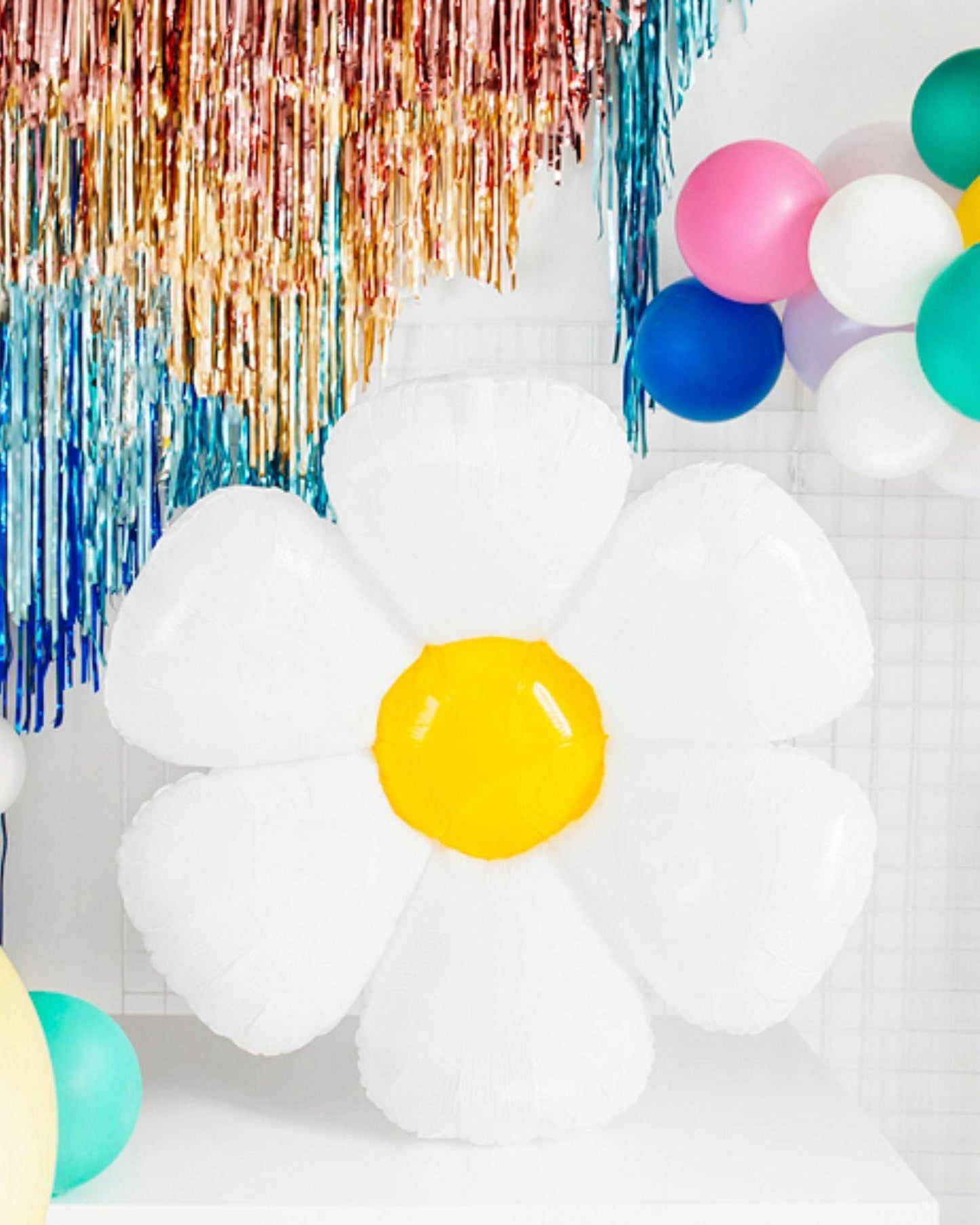 A large white flower with a yellow center, surrounded by colorful balloons and shiny decorative streamers.