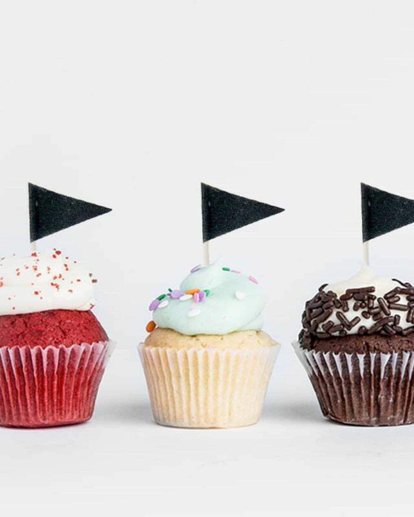 Three colorful cupcakes topped with various icing and small black flags, arranged on a light background.
