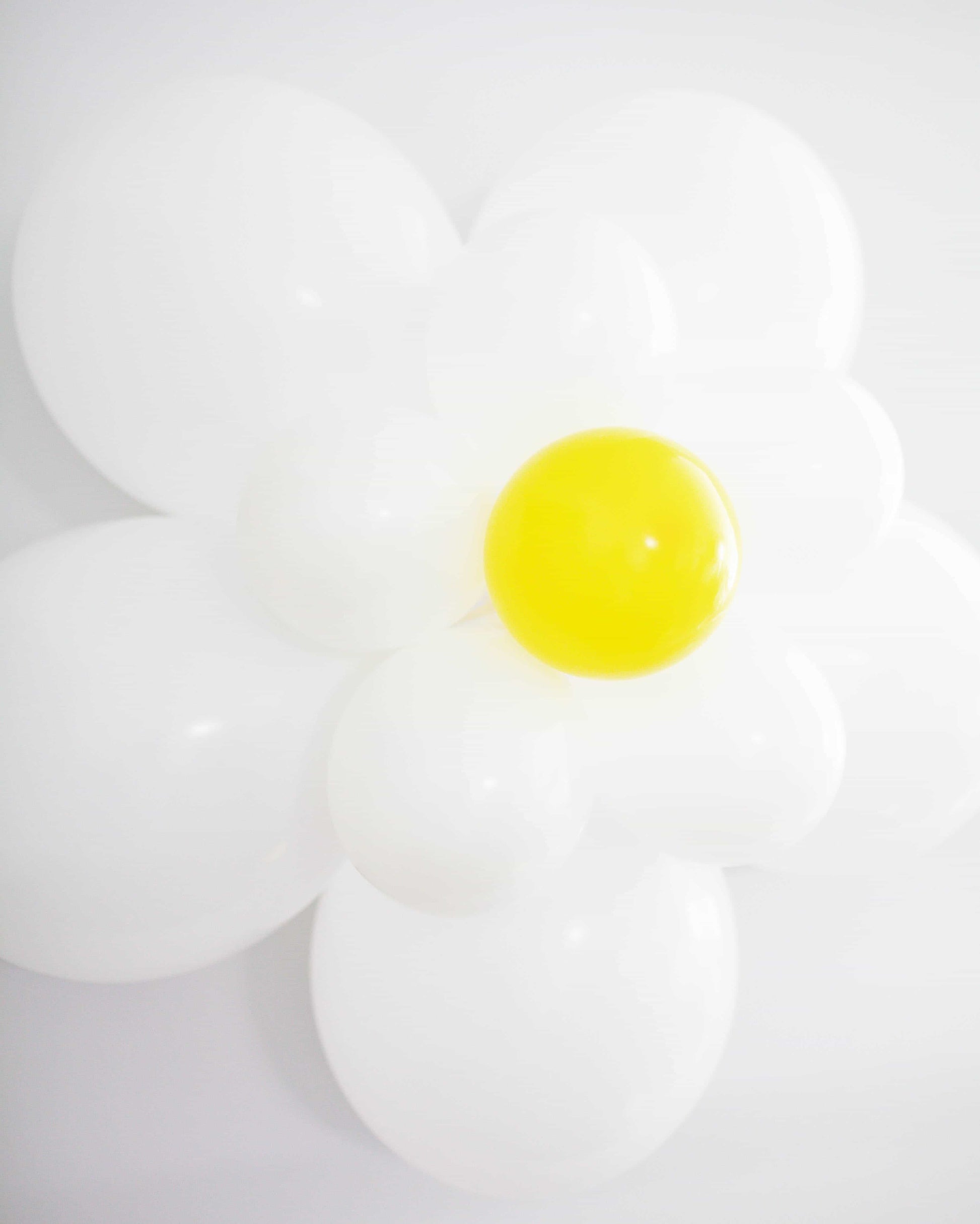 A cluster of white balloons forms a flower shape, with a bright yellow center, against a light backdrop.