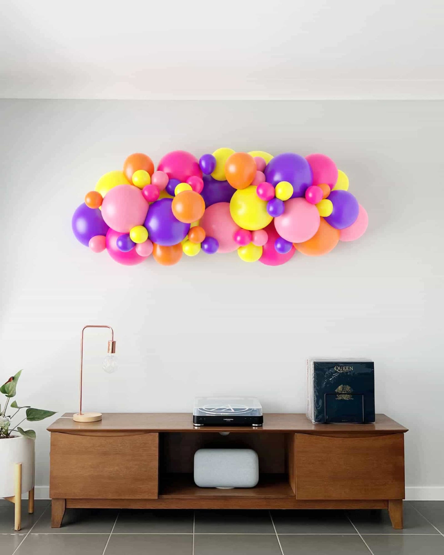 Colorful balloon garland in pink, purple, yellow, and orange, above a wooden console table with decor and a plant.