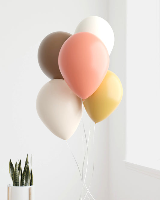 A cluster of pastel balloons in soft colors, gently floating beside a small potted plant against a light wall.