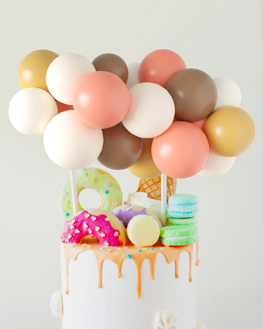 Colorful balloons hover above a cake adorned with dripping icing, donuts, macarons, and an ice cream cone.