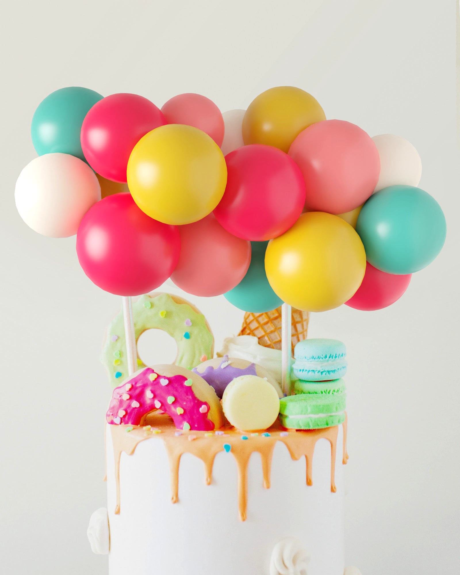 Colorful balloons hover above a cake adorned with vibrant sweets, including donuts, macarons, and an ice cream cone.