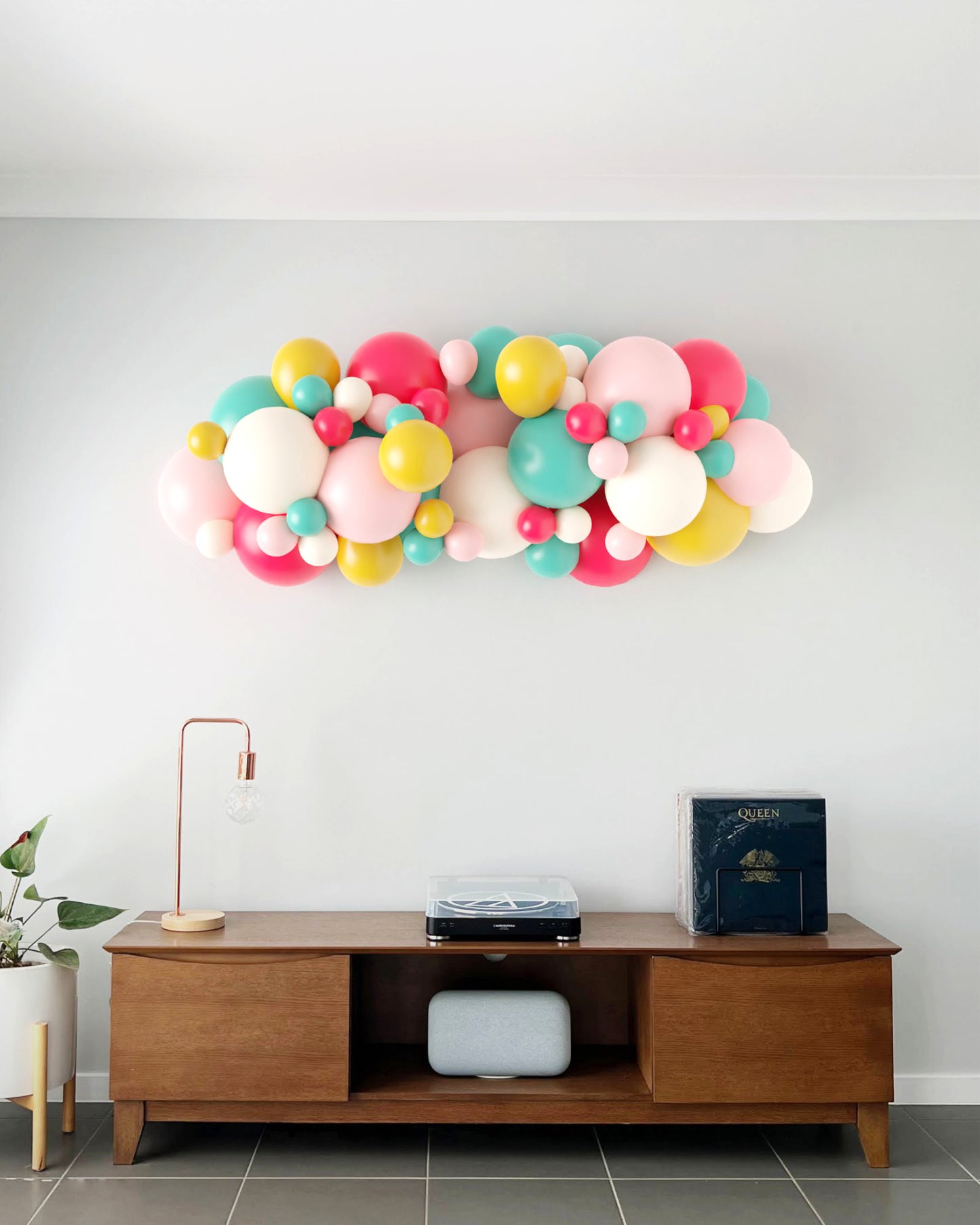 Colorful balloon garland in pink, yellow, teal, and red, adorning a light wall above a wooden table with decor.