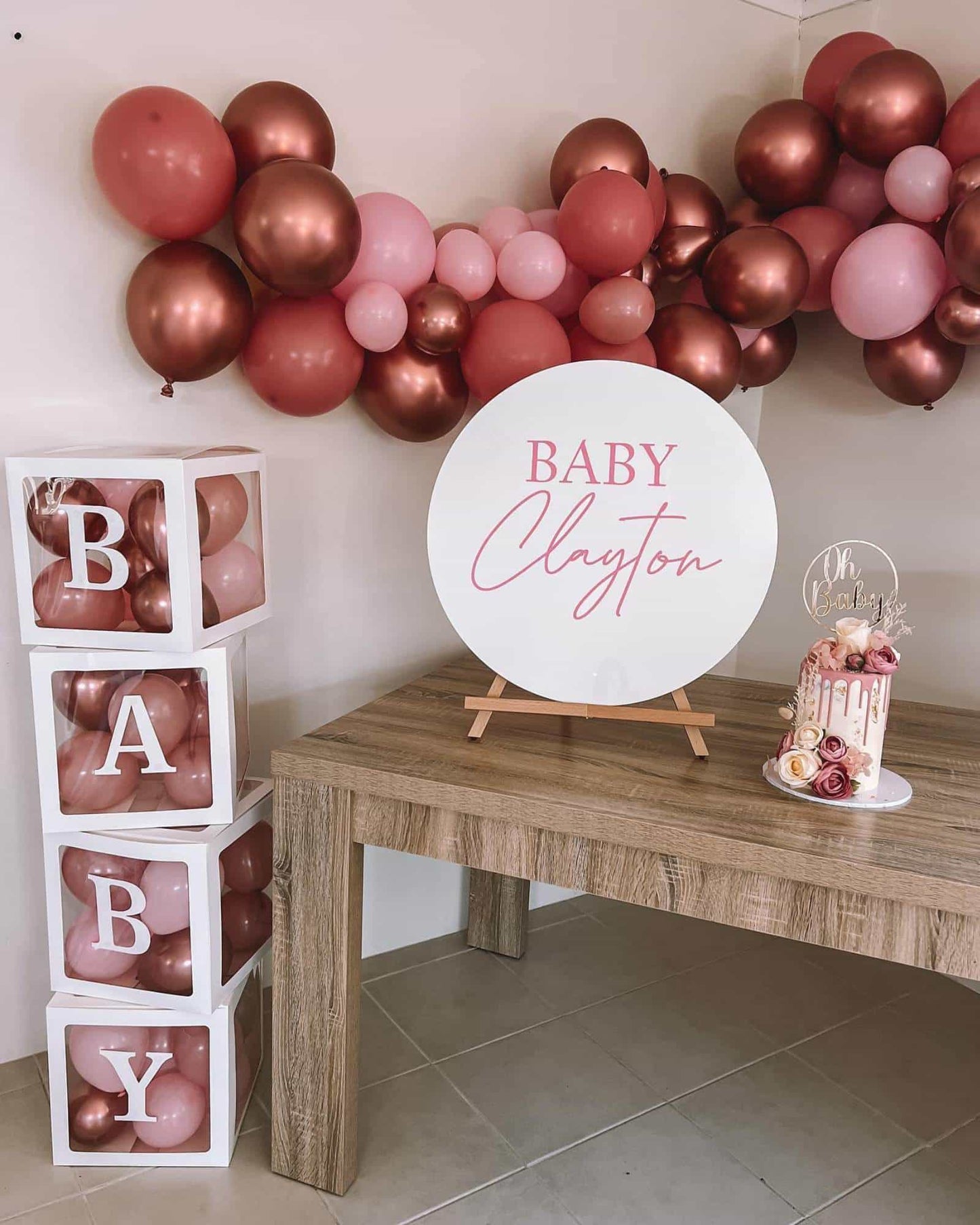 A festive display with pink and copper balloons, a sign reading "Baby Clayton," and a decorative cake on a wooden table.