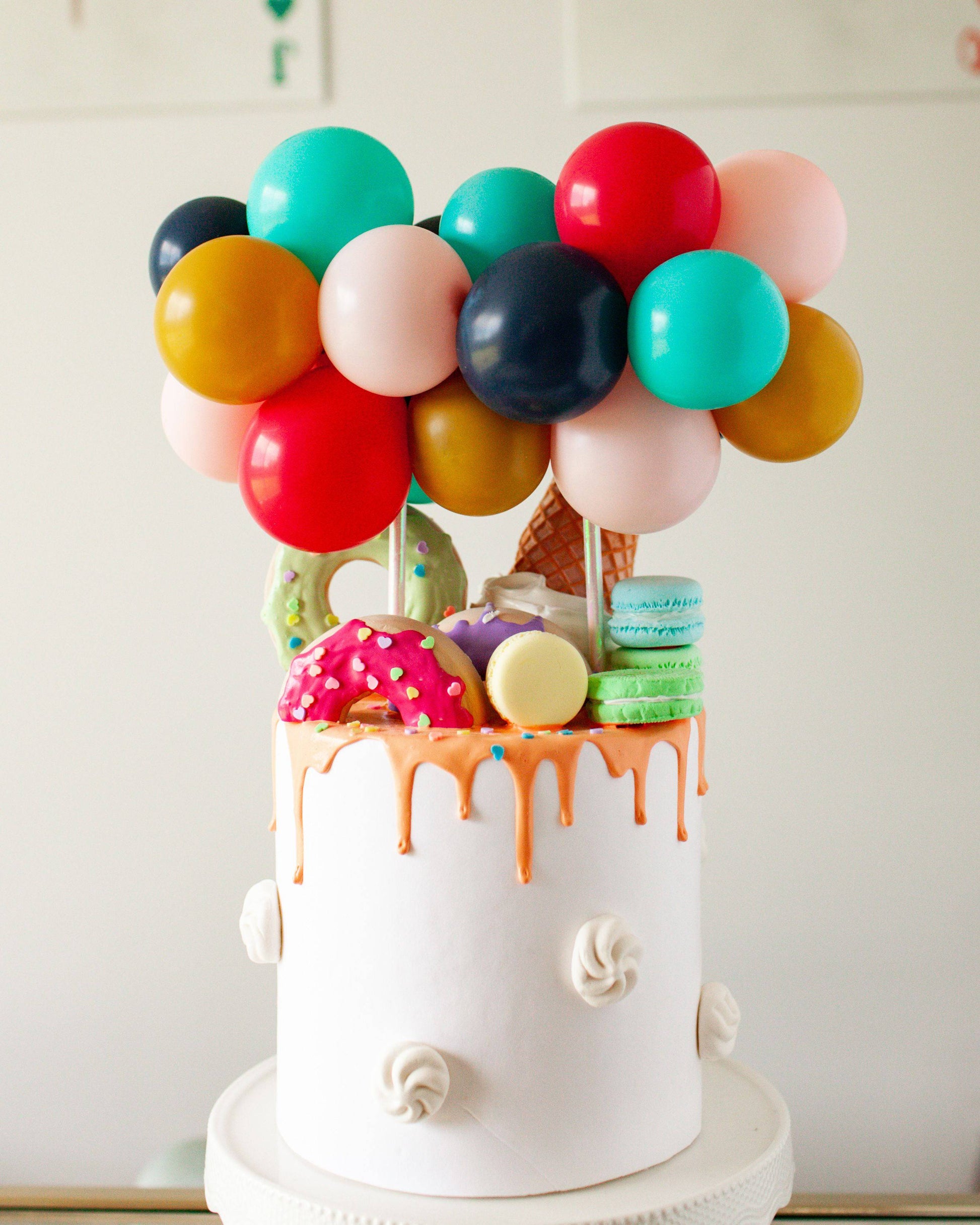 A vibrant cake topped with colorful balloons and playful sweets, featuring drips of orange icing and decorative accents.