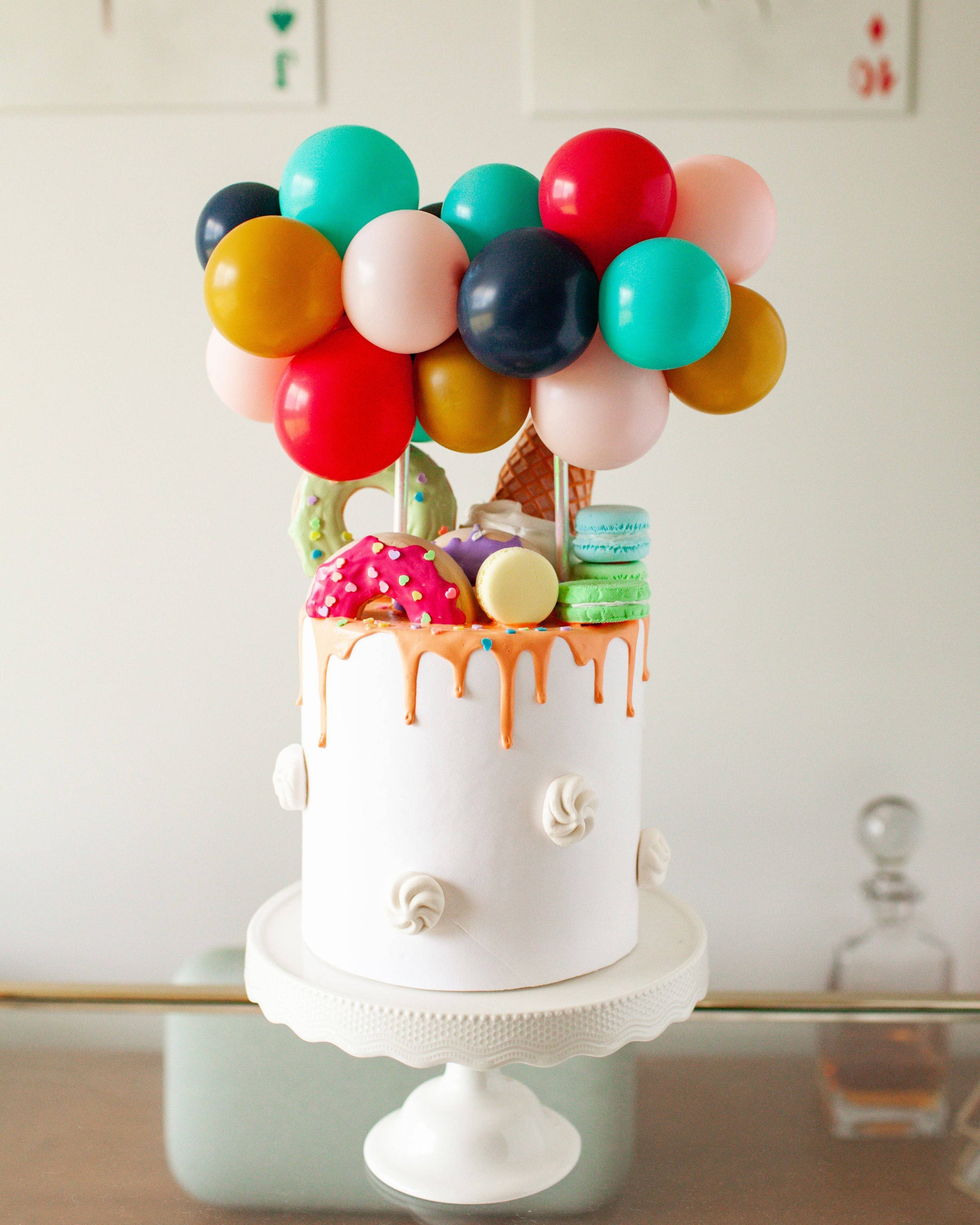 A whimsical cake topped with colorful balloons, sprinkles, and various sweet treats, set on a decorative stand.