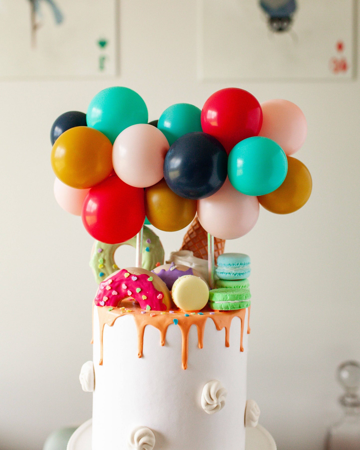 Colorful balloons top a white cake drizzled with orange, adorned with donuts, macarons, and an ice cream cone.