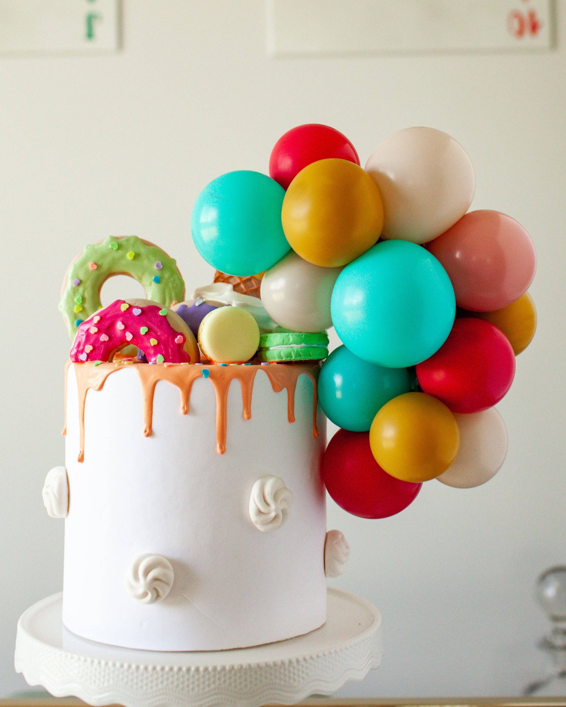 Colorful balloons and sweet treats adorn a white cake with dripping icing, creating a festive and vibrant display.