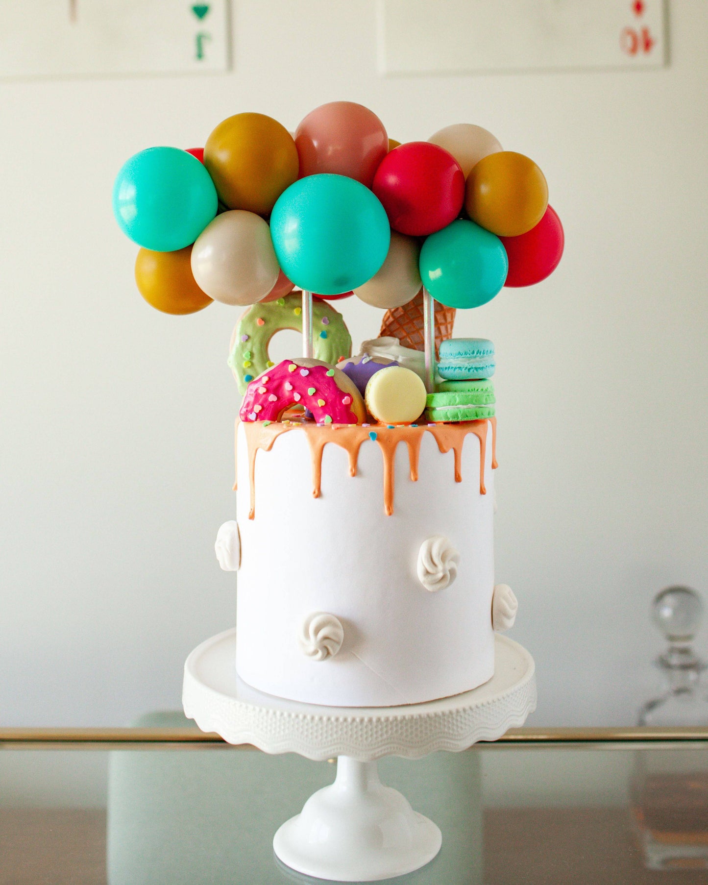 Colorful balloons atop a white cake adorned with dripping icing, sprinkles, and various sweet treats.