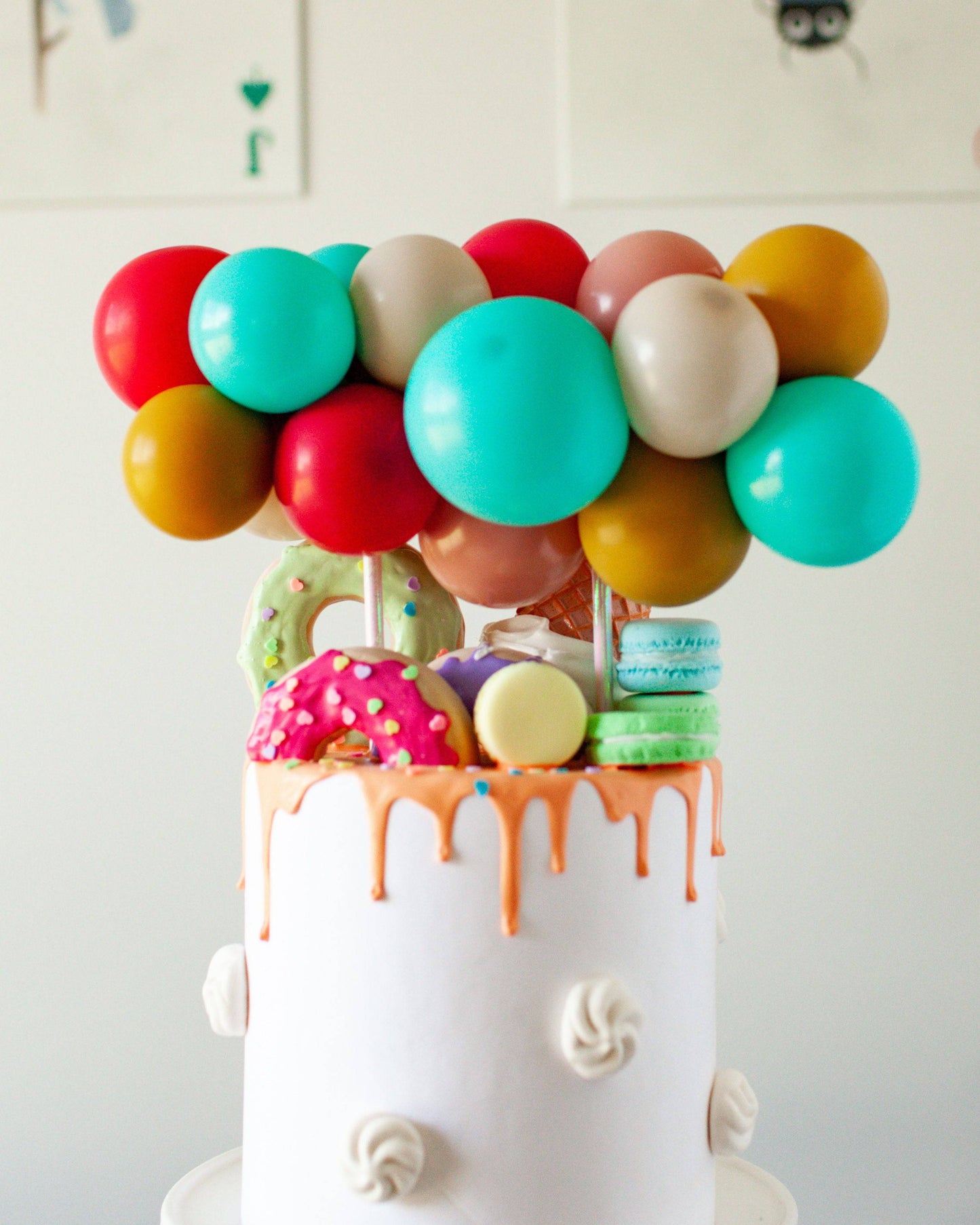 Colorful balloons top a white cake adorned with dripping icing and vibrant confections, creating a festive display.
