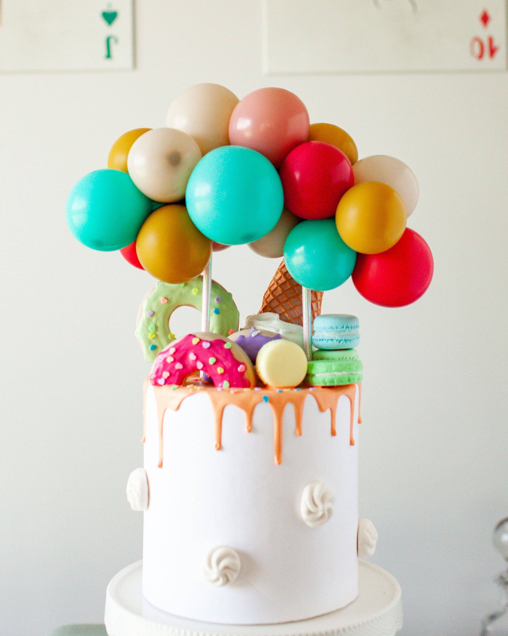 A colorful cake topped with balloons, donuts, ice cream, and macarons, featuring vibrant sprinkles and drips.
