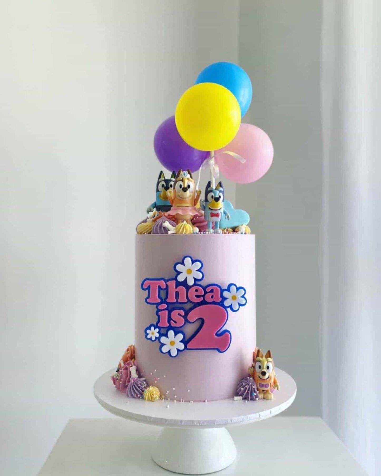 A pastel purple cake adorned with colorful balloons and playful animal figures, celebrating a second birthday.
