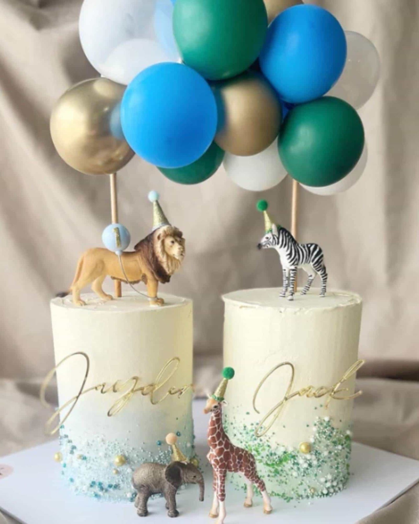 Two layered cakes topped with colorful balloons and playful animal figurines, featuring gold lettering for names.