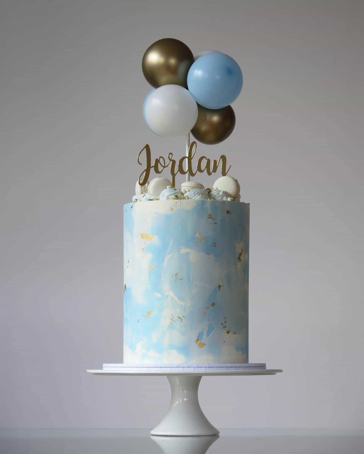 A blue cake adorned with golden accents, topped with balloons and a name in gold script, on a white pedestal.
